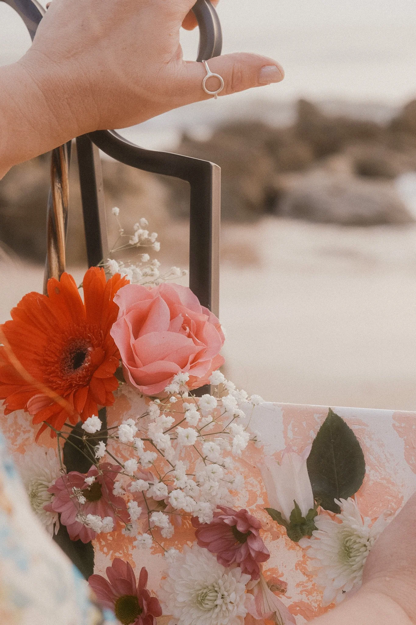 Close-up of a person's hand holding a flower arrangement with pink roses, white baby's breath, orange daisies, and greenery on a pink and white patterned surface.