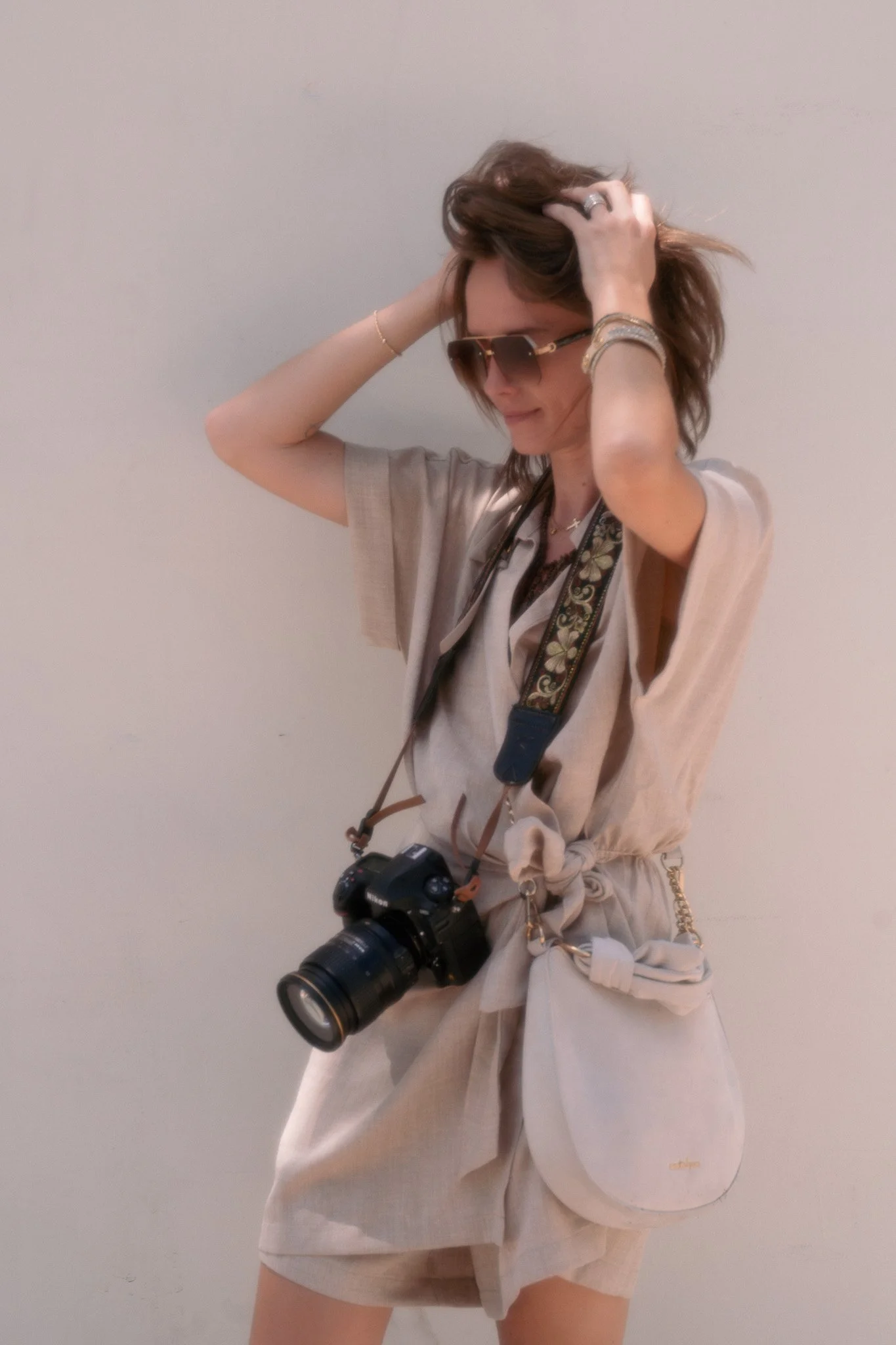 A woman wearing sunglasses and a beige outfit, holding her hair with both hands, with a large professional camera hanging around her neck, standing against a plain white wall.