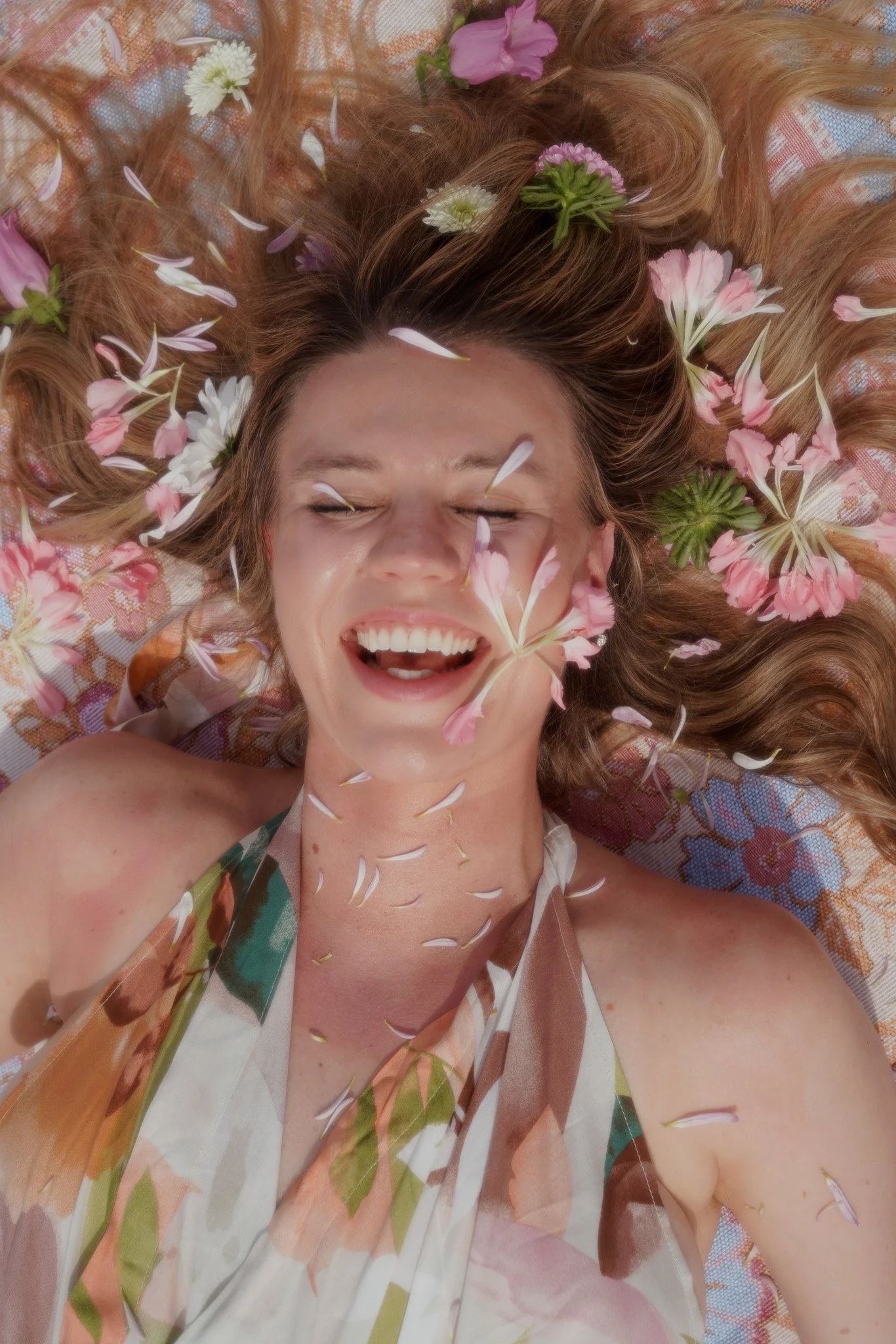 A woman with long auburn hair lying down with her eyes closed and mouth open in a smile, surrounded by pink, white, and purple flowers and petals on her hair and a patterned cloth underneath.