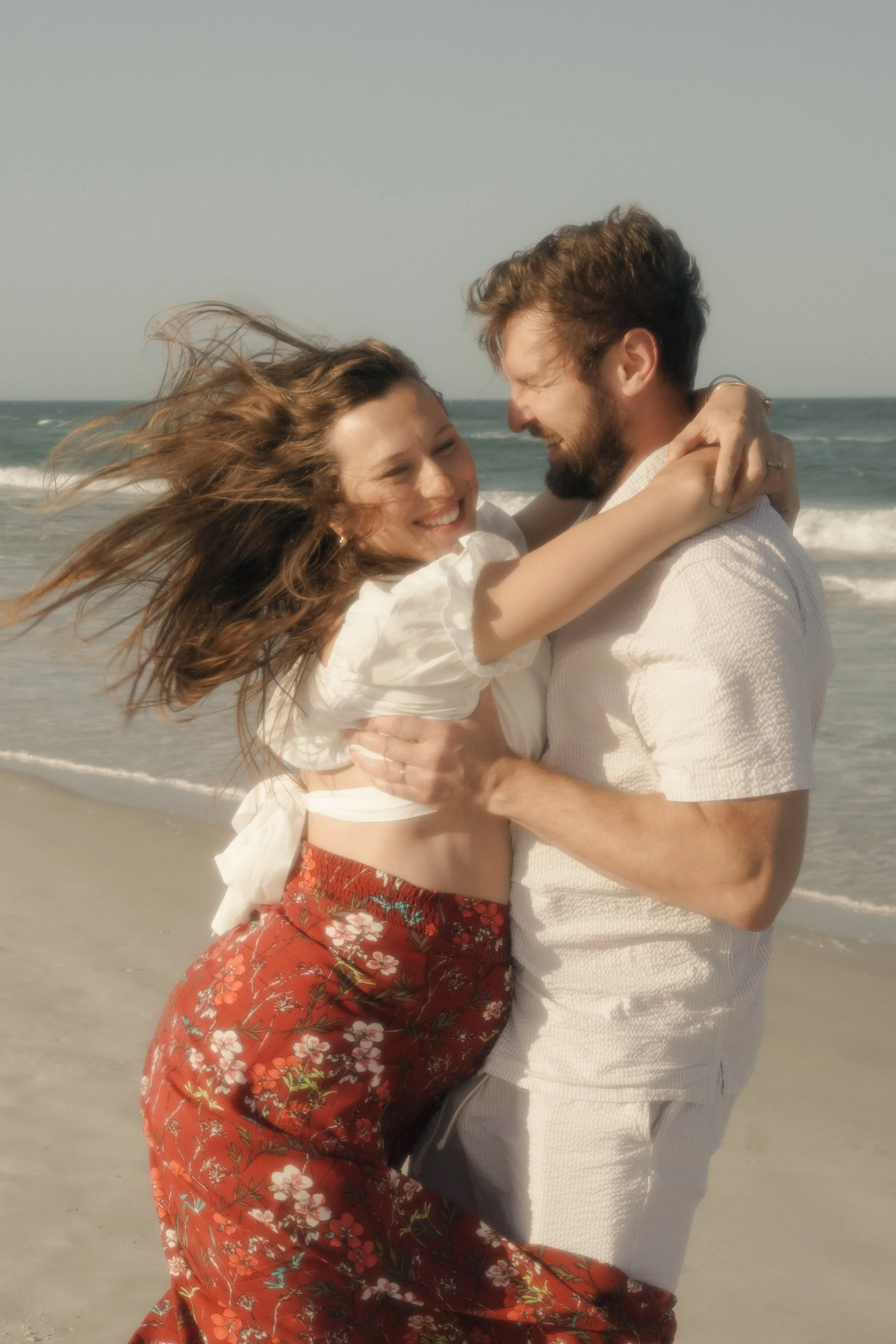 A smiling man and woman at the beach, embracing each other with the ocean and sky in the background.