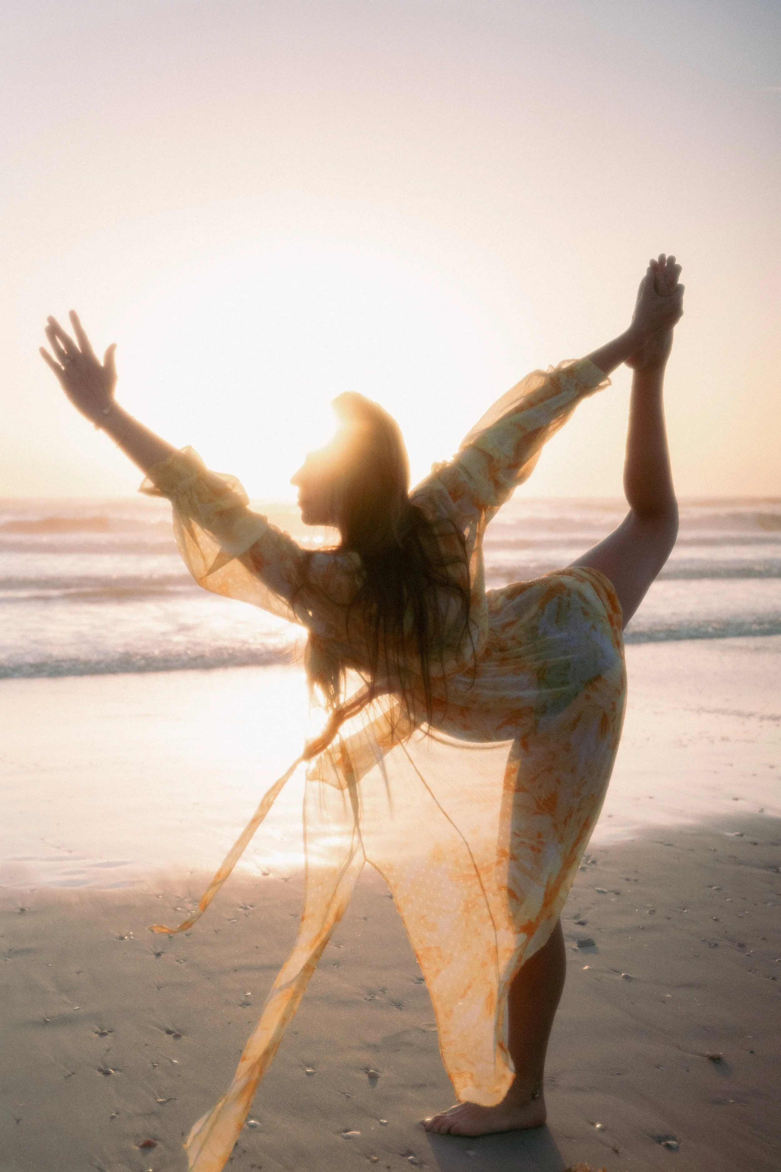 A woman practicing yoga on the beach at sunset, standing on one leg with arms raised and hands in prayer position.