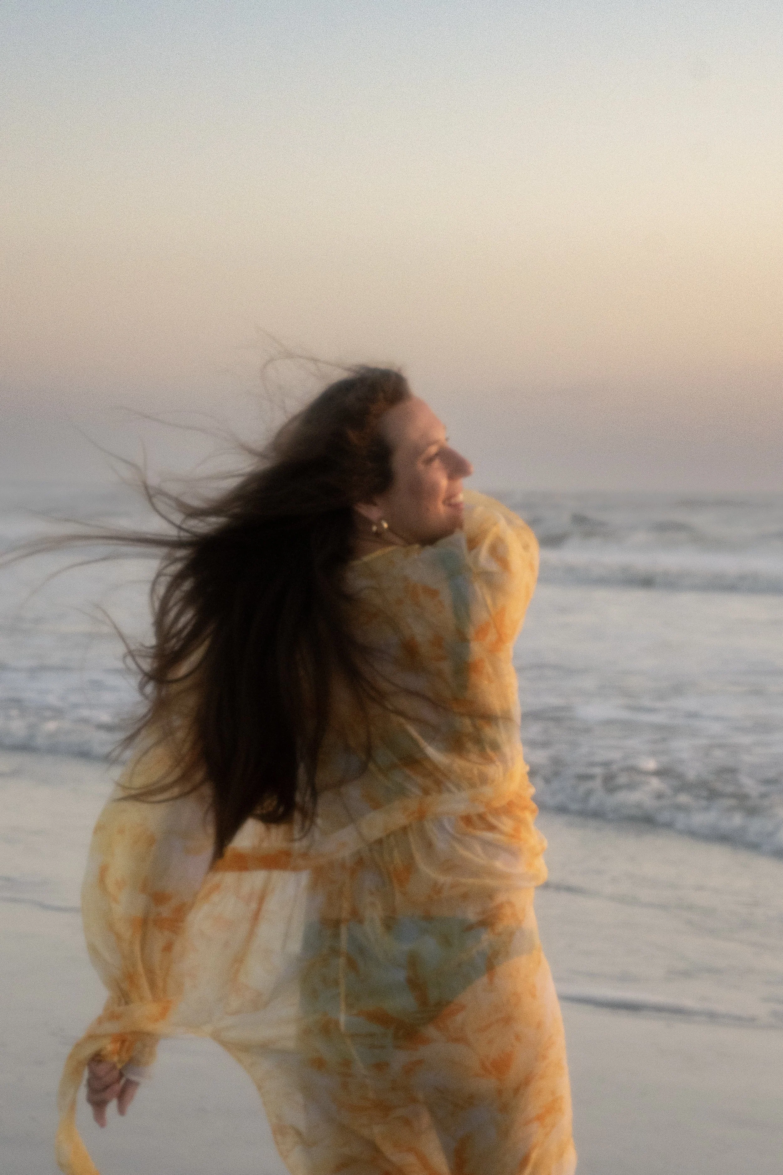 A woman with long dark hair wearing a yellow and orange tie-dye hoodie, smiling and looking to the side, standing on a beach with ocean waves and a pastel-colored sky in the background.