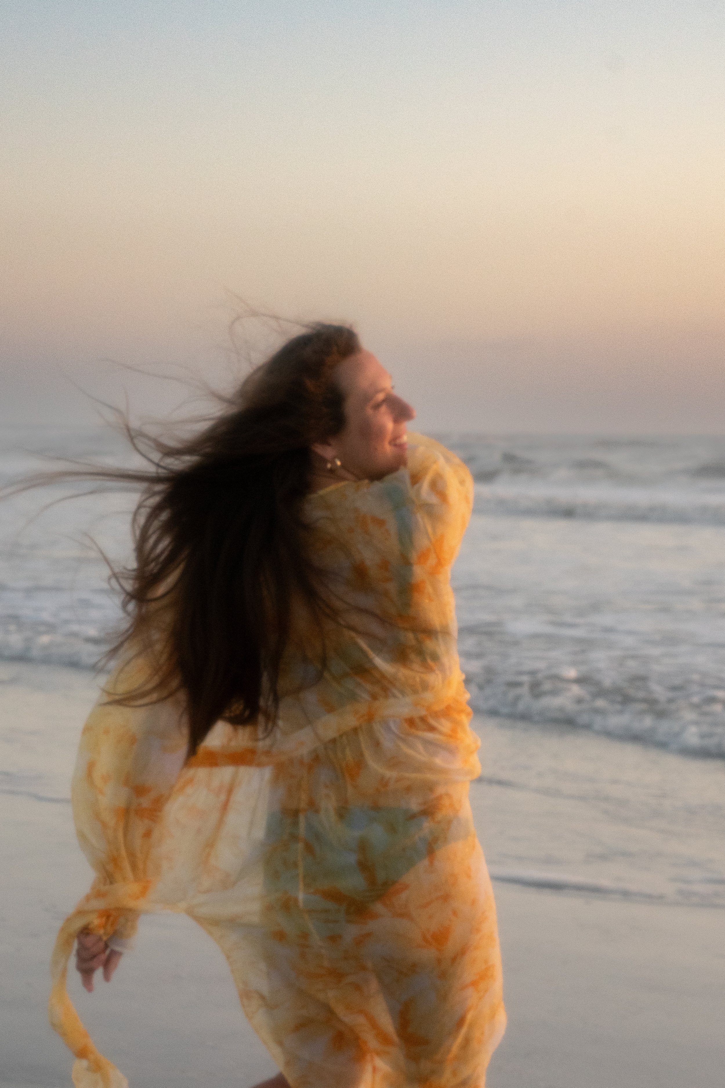 A woman with long, dark hair stands on the beach at sunset, smiling as she faces the ocean, wearing a yellow and orange tie-dye dress.