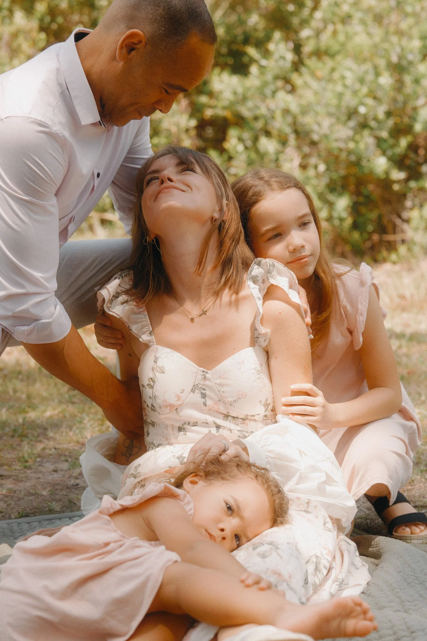 A man and two young girls relaxing outdoors in a park. The man is smiling and leaning toward one girl, while the other girl is sitting next to him, looking at the camera with a neutral expression. One girl is lying on the ground, looking at the camera. All are dressed in light-colored clothing, with trees and natural scenery in the background.