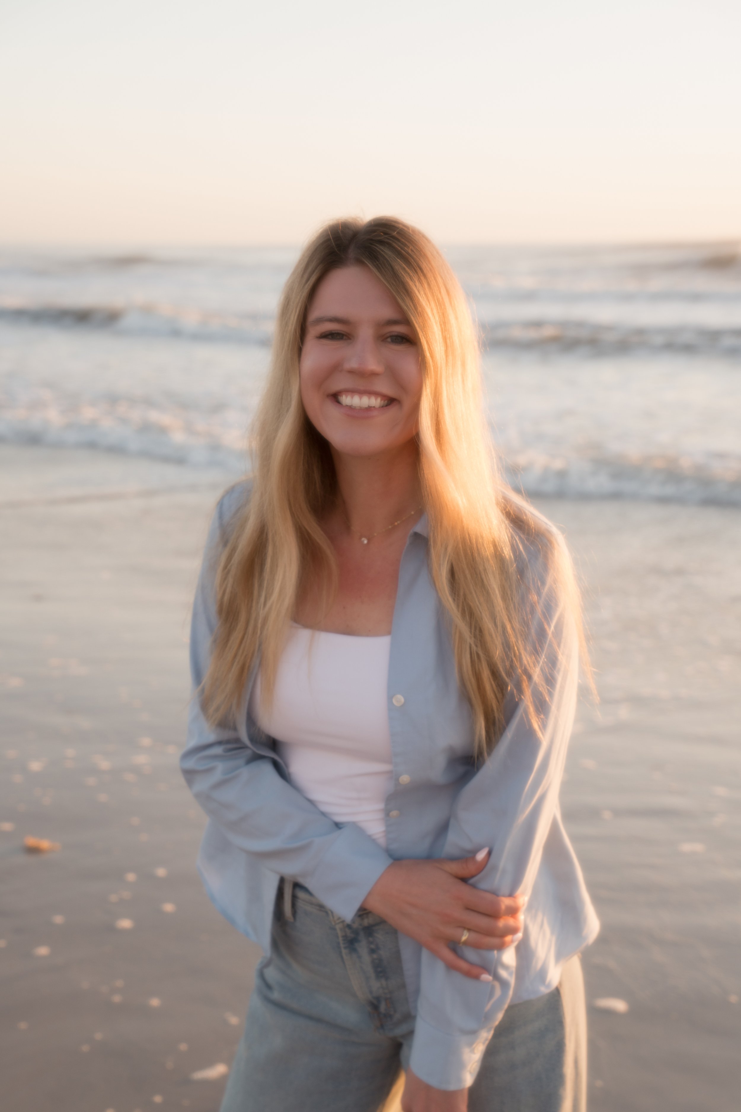 A woman smiling at the beach during sunset, wearing a light blue jacket, white top, and gray pants with waves in the background.