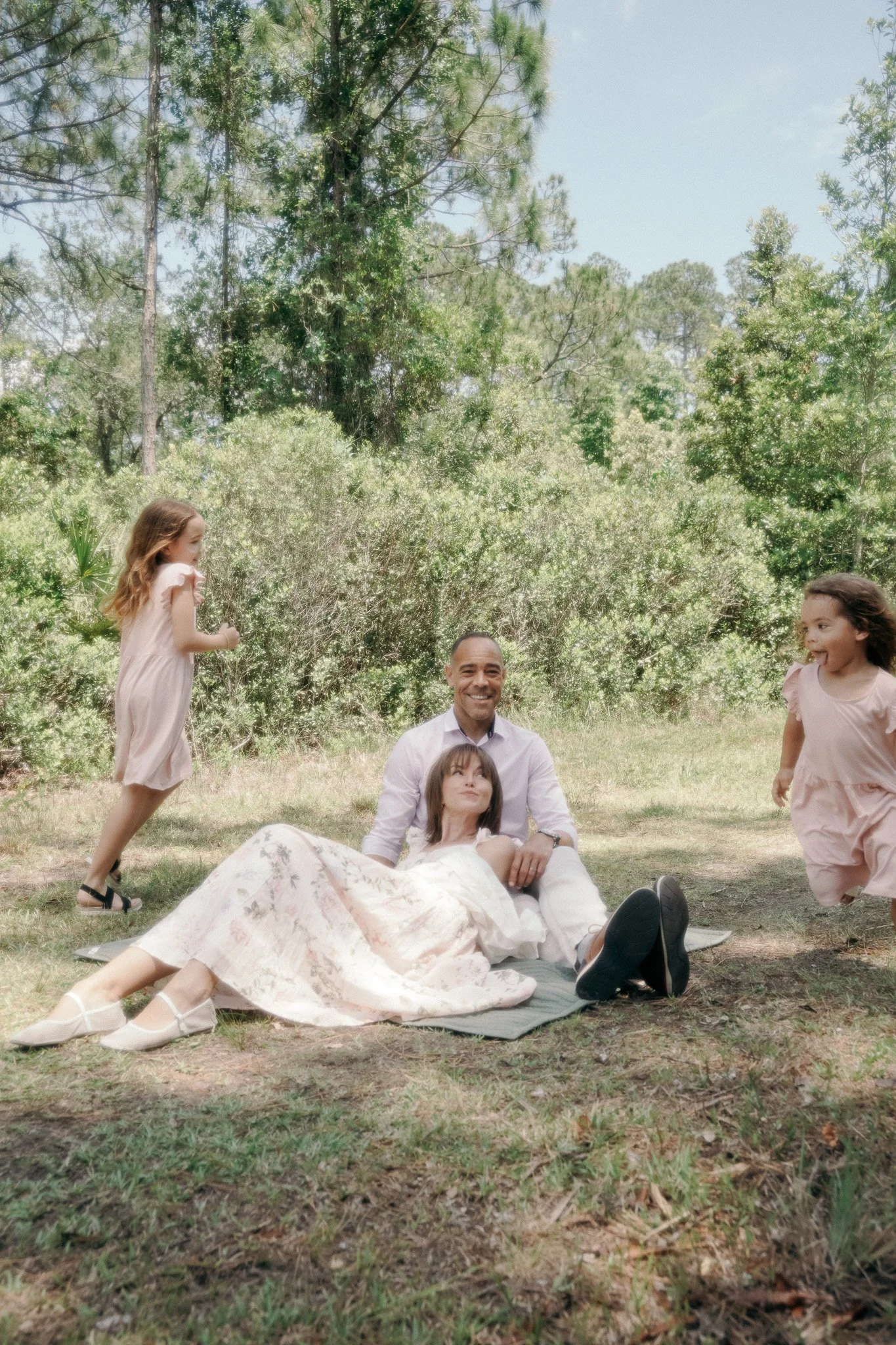 A man and woman lying on the grass in a forested area, with three young girls playing around them, all dressed in light-colored clothing on a sunny day.