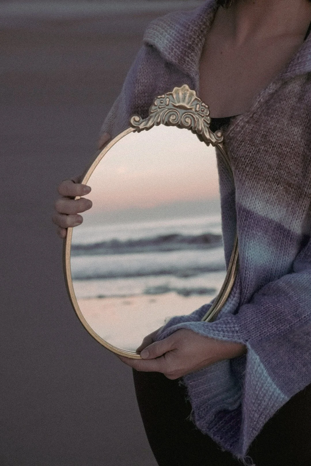 Person holding an ornate round mirror reflecting a beach and sunset scene.