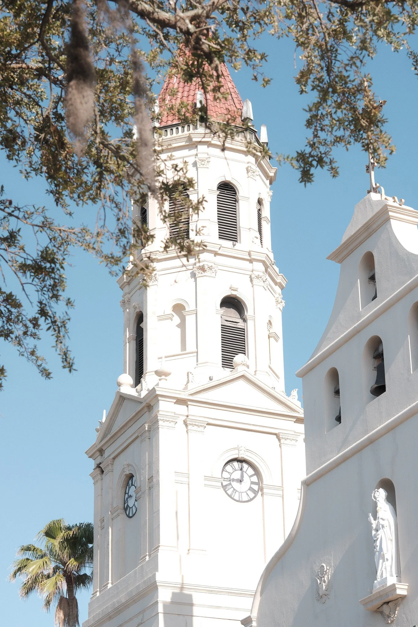 A white clock tower with a red-tiled roof, surrounded by trees and a white building with decorative statues and architectural details, under a clear blue sky.