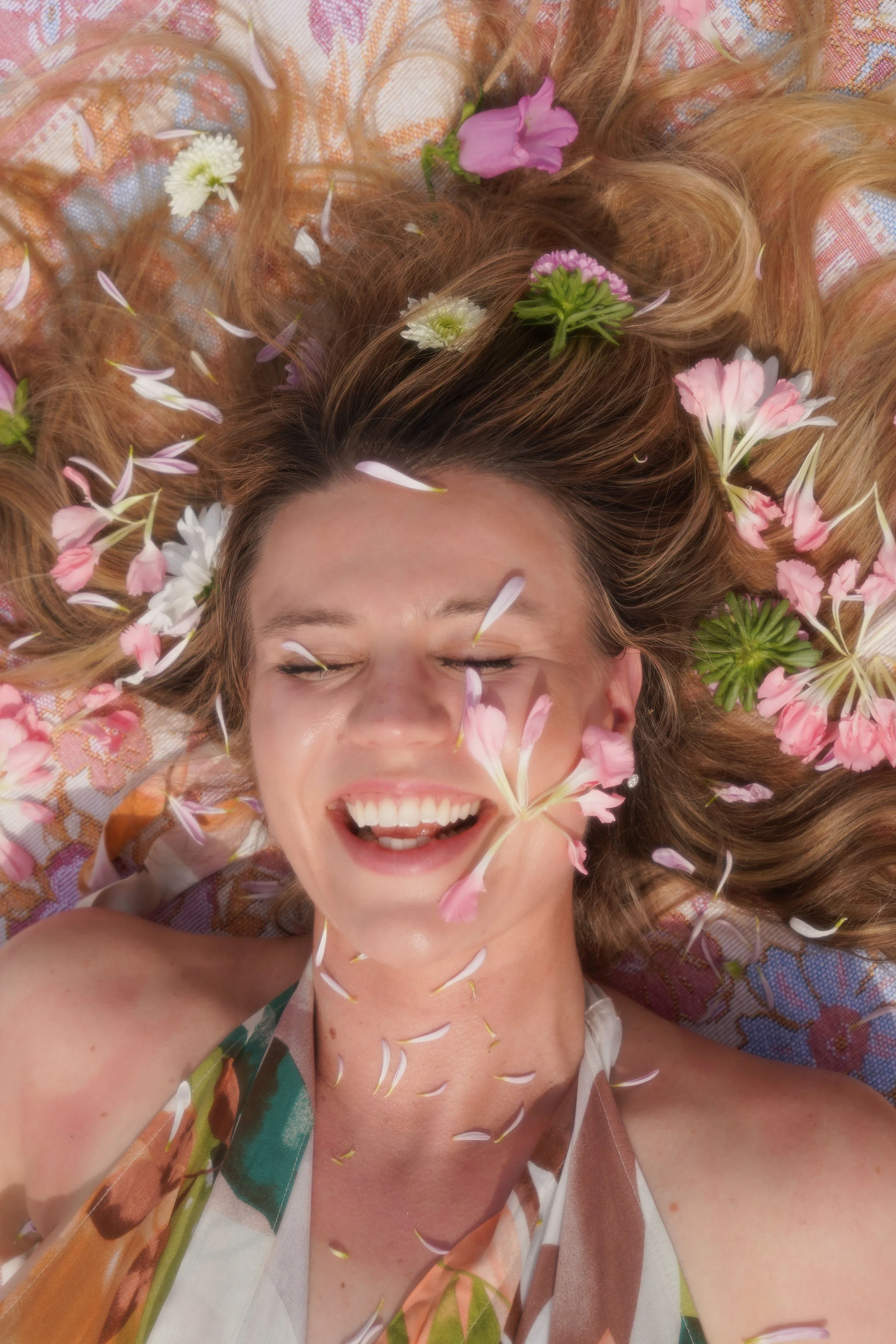 A woman lying down with long, wavy hair, smiling with her eyes closed, surrounded by pink and white flowers and flower petals.