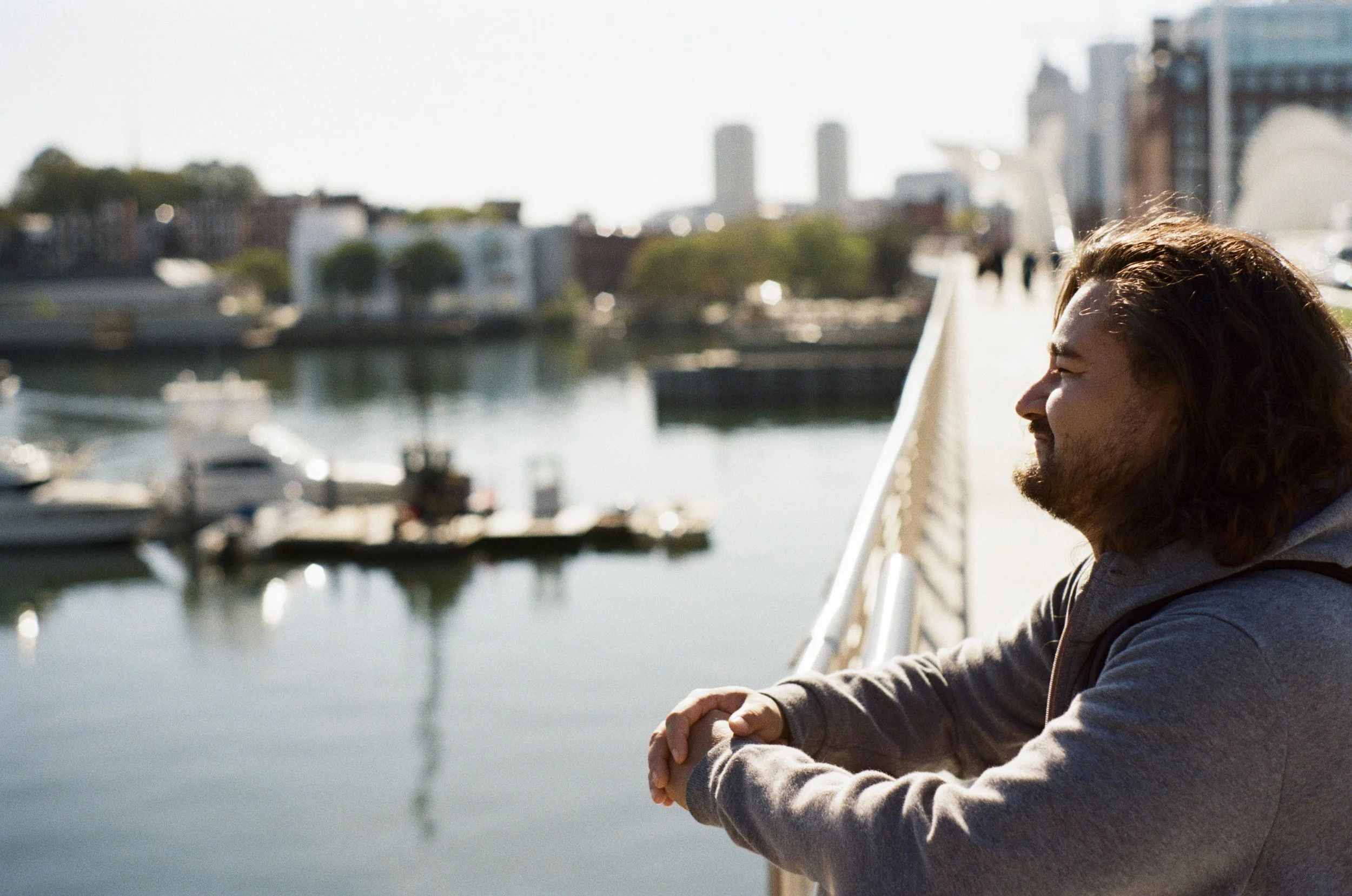 A young man with long hair and a beard is sitting outdoors near a body of water on a bridge, smiling with his eyes closed, wearing a gray hoodie.