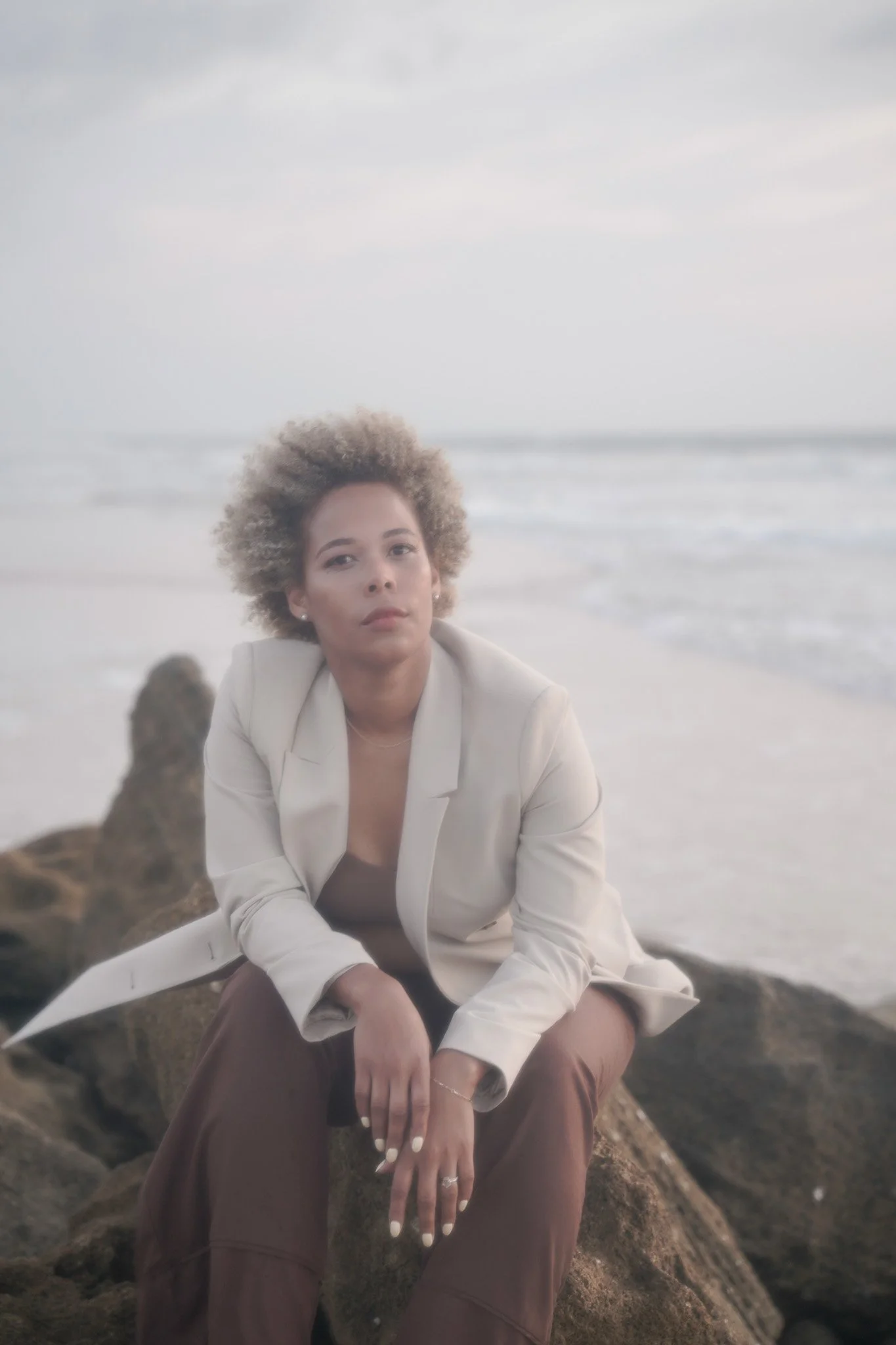 A woman with curly blonde hair sitting on rocks at the beach during overcast weather, wearing a white blazer and brown pants.
