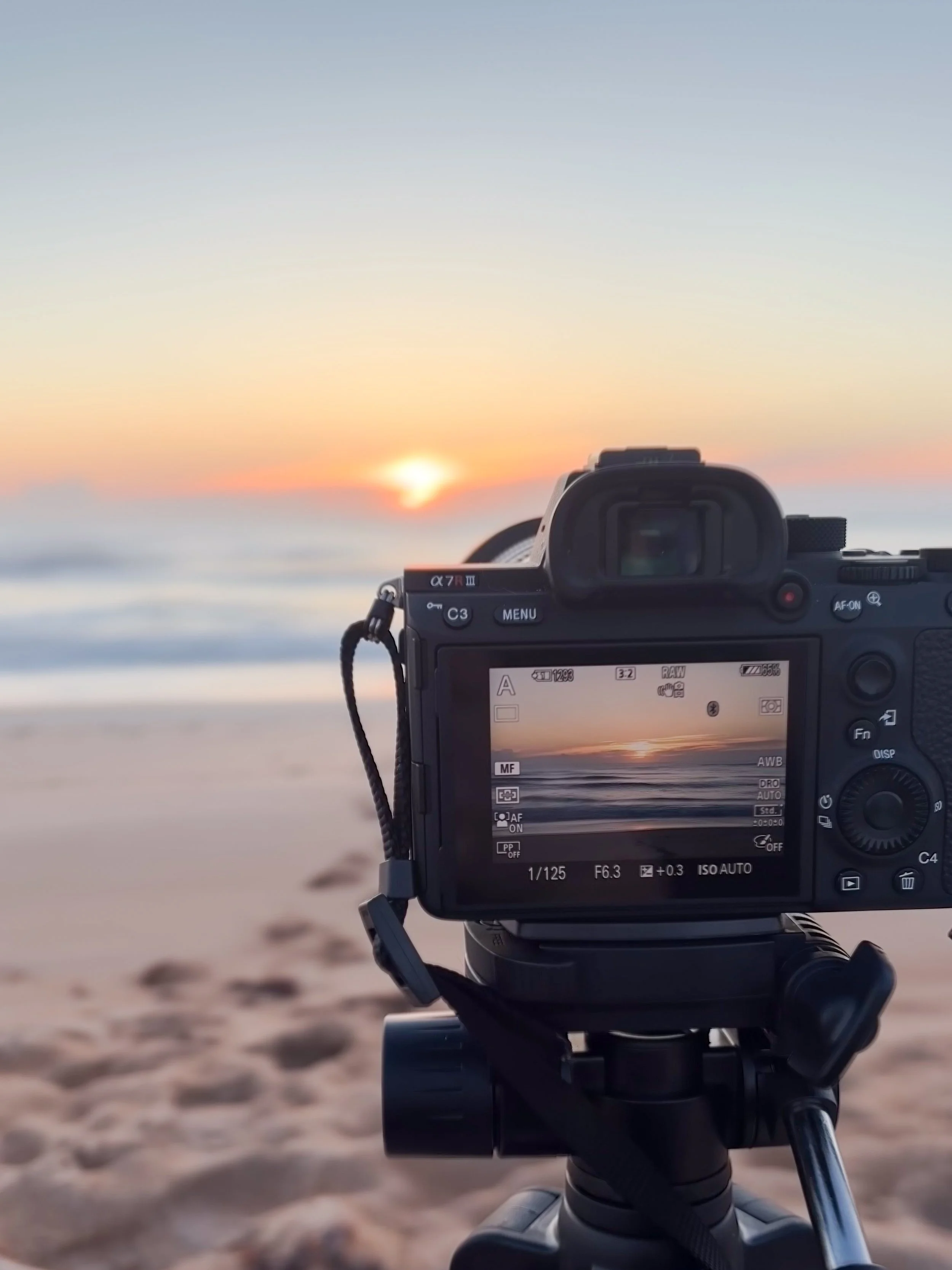 A digital camera mounted on a tripod captures a sunset over the ocean with a sandy beach in the foreground.