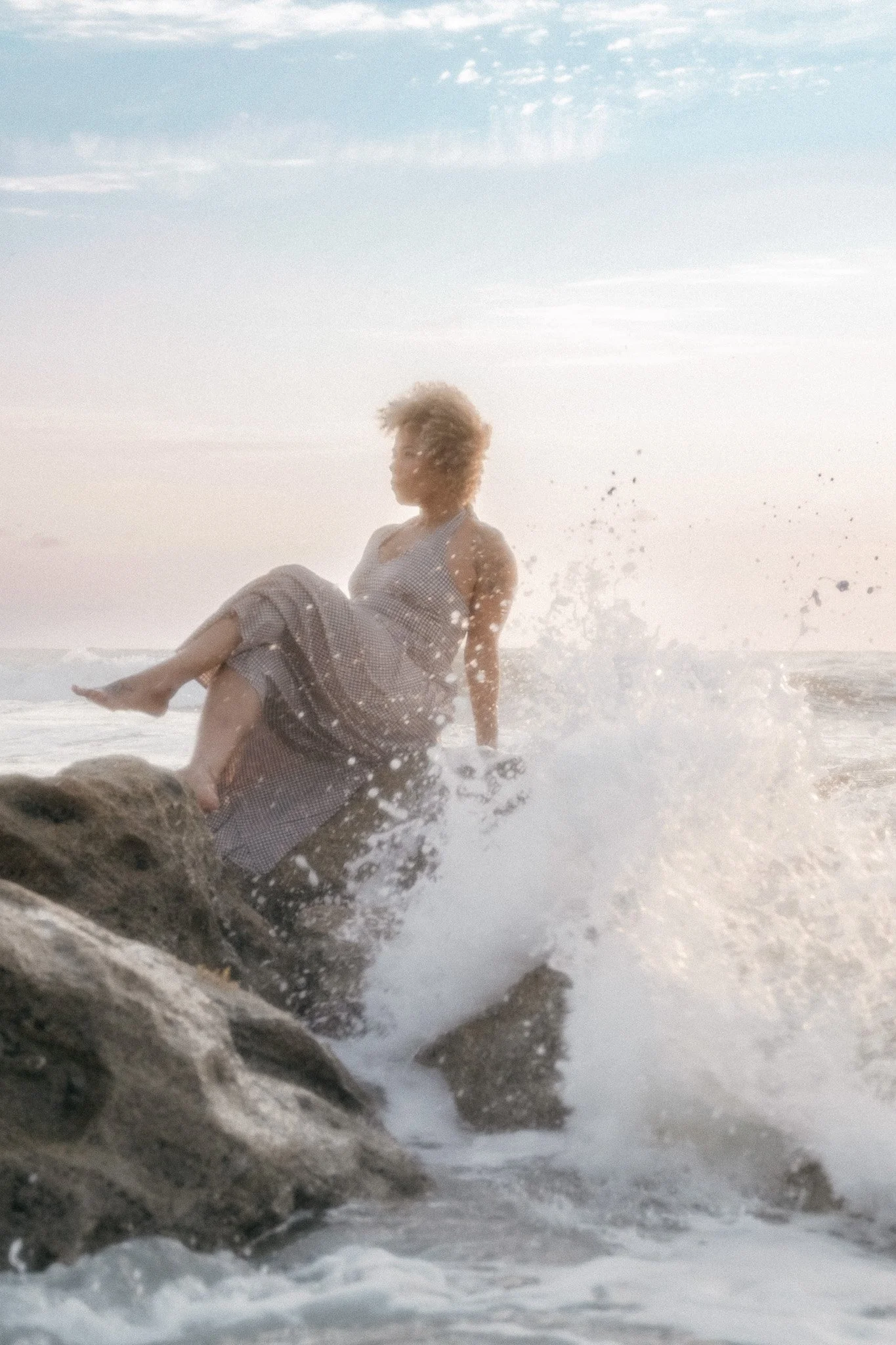 A woman with curly hair sitting on rocks near the ocean, with waves splashing around her during sunset or sunrise.