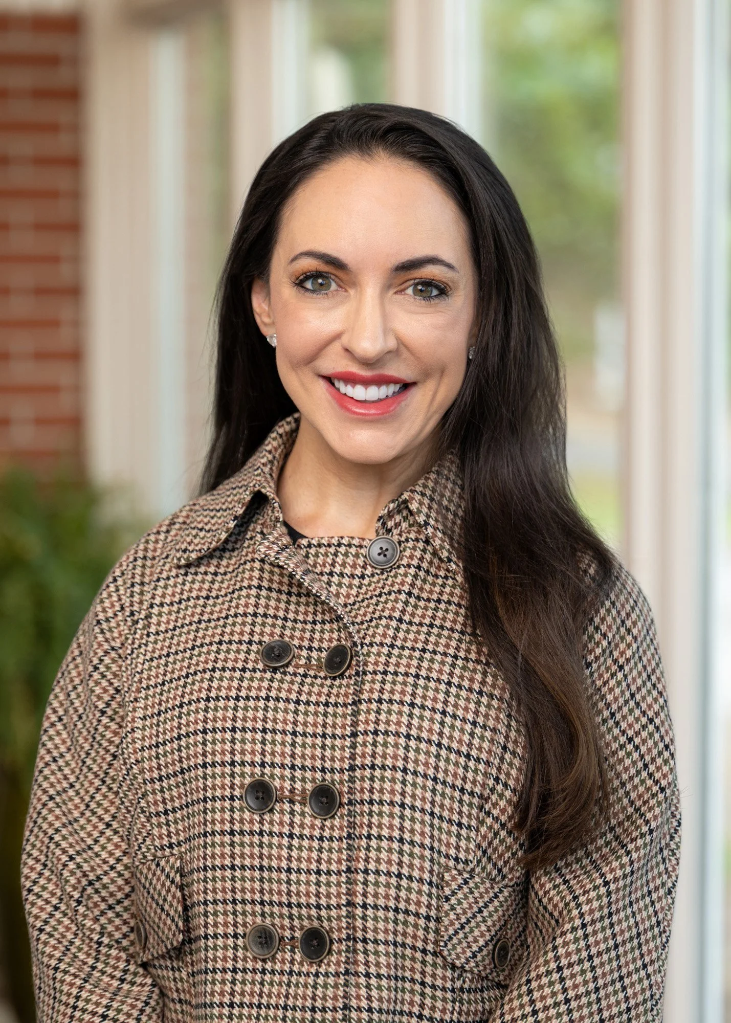 A woman with long dark hair, smiling, wearing a plaid coat with large buttons, standing inside near large windows with a view of trees outside.