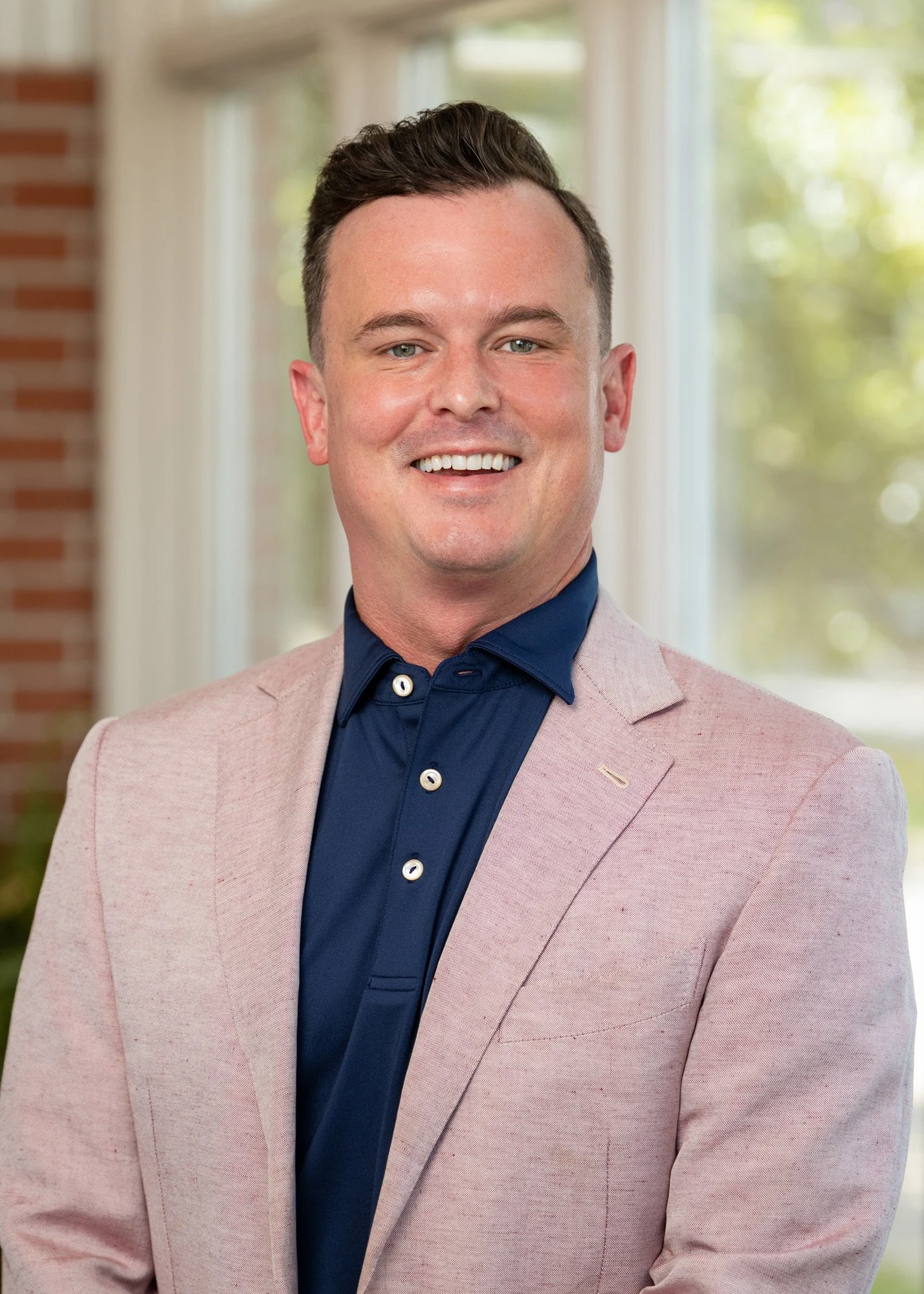 A smiling man in a light pink blazer and navy blue shirt, standing in front of a window with trees outside.