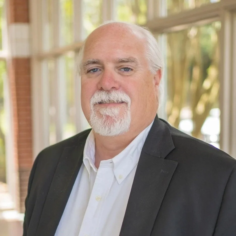 A middle-aged man with white hair and a beard wearing a black suit jacket and white shirt, standing indoors with large windows and greenery outside.