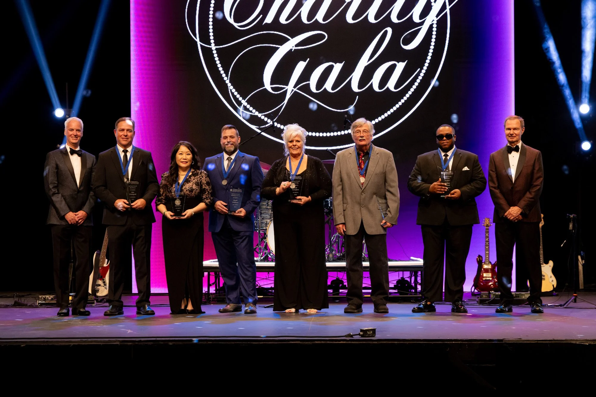 Group of seven people standing on stage at Charity Gala, some holding awards, backdrop displays 'Charity Gala', stage lights, musical instruments in background.