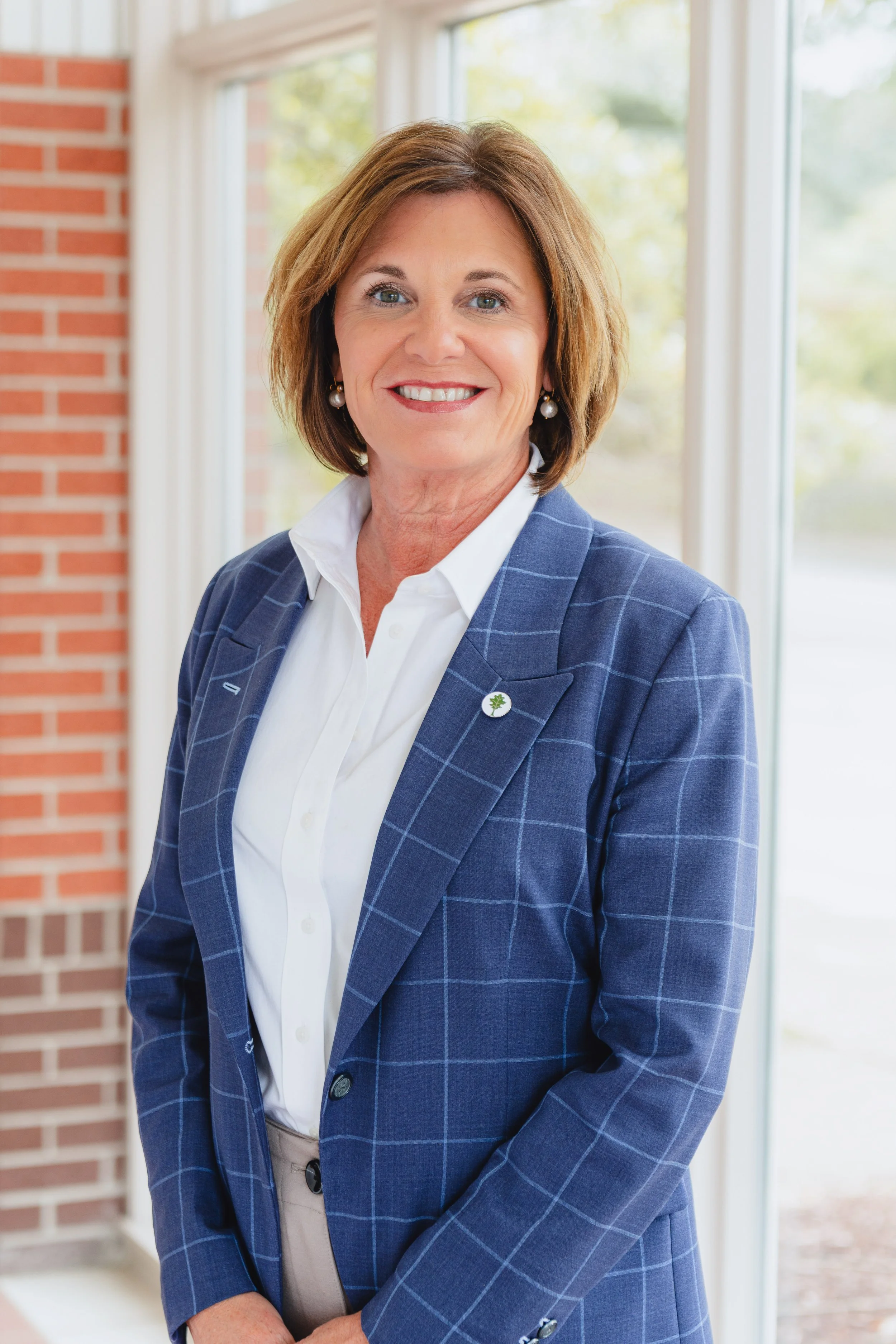 A smiling woman with shoulder length light brown hair wearing a white shirt and a blue checkered blazer with earrings and a pin, standing in front of a large window showing trees outside, brick wall on the side.