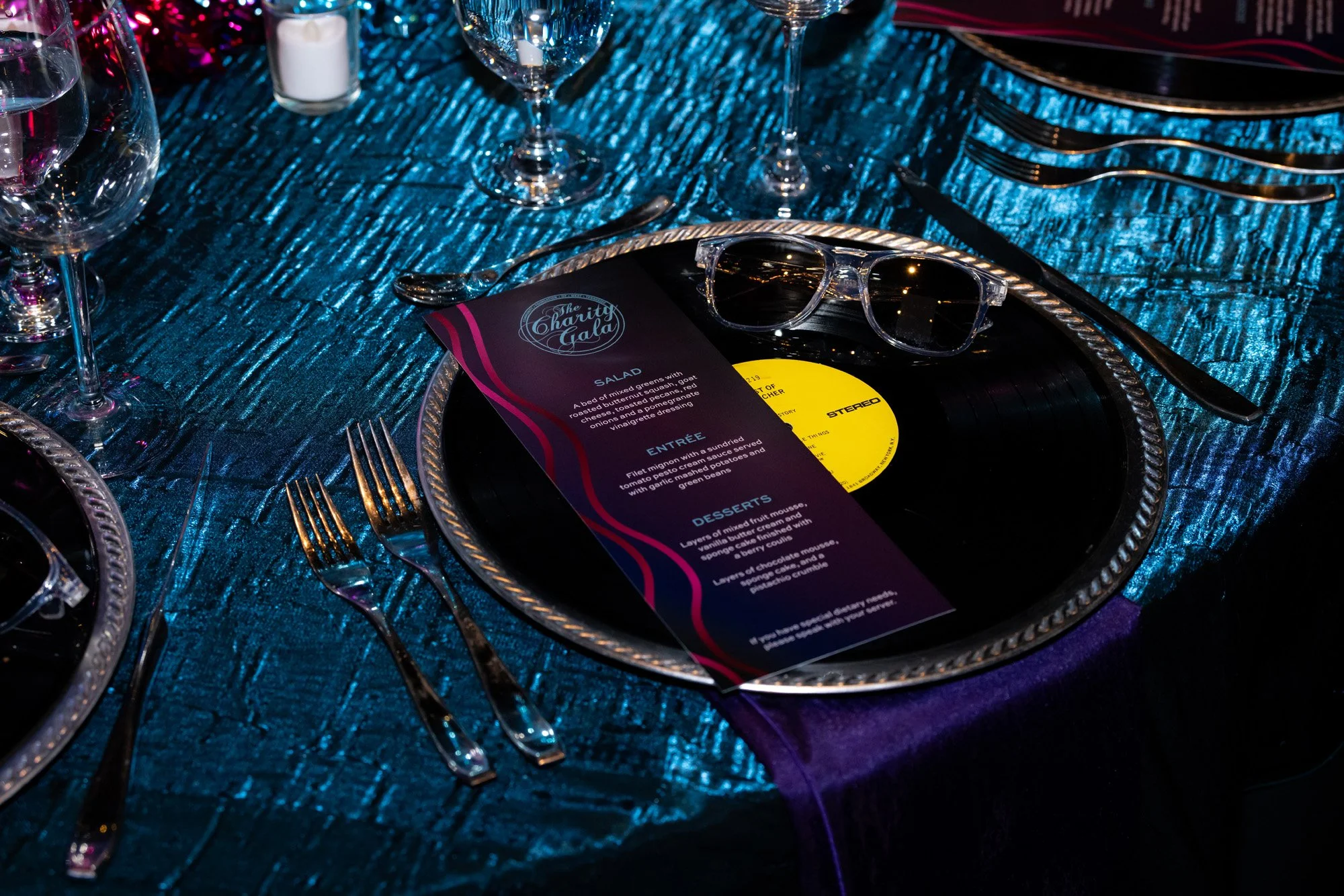 Table set at a charity gala with a violet-blue textured tablecloth, wine glasses, cutlery, a candle, and sunglasses on a plate holding the event's menu.