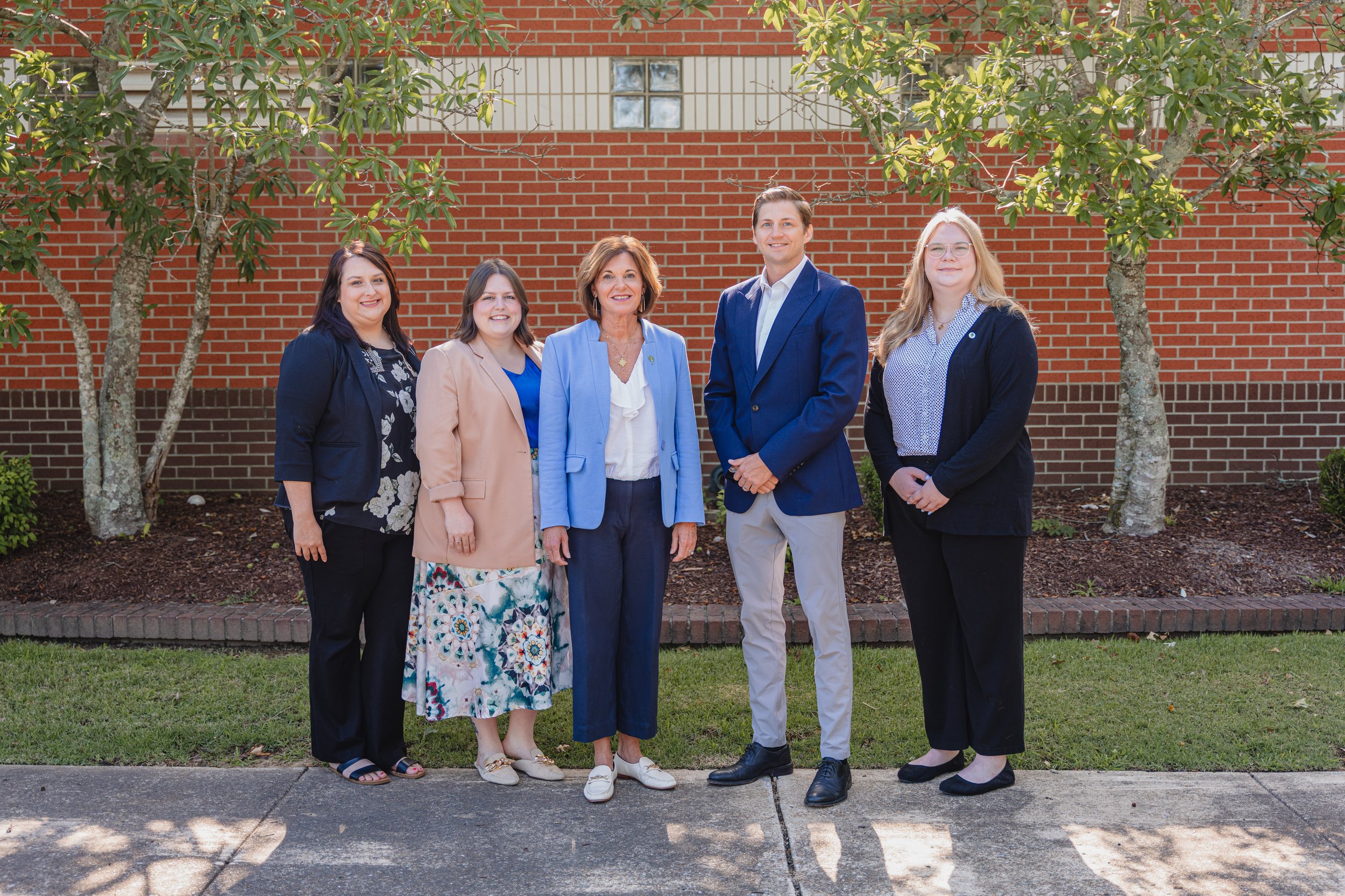 Community Foundation team members standing outdoors in front of a brick wall and green trees, posing for a group photo. The group includes five women and one man, all dressed in business casual attire, smiling at the camera.