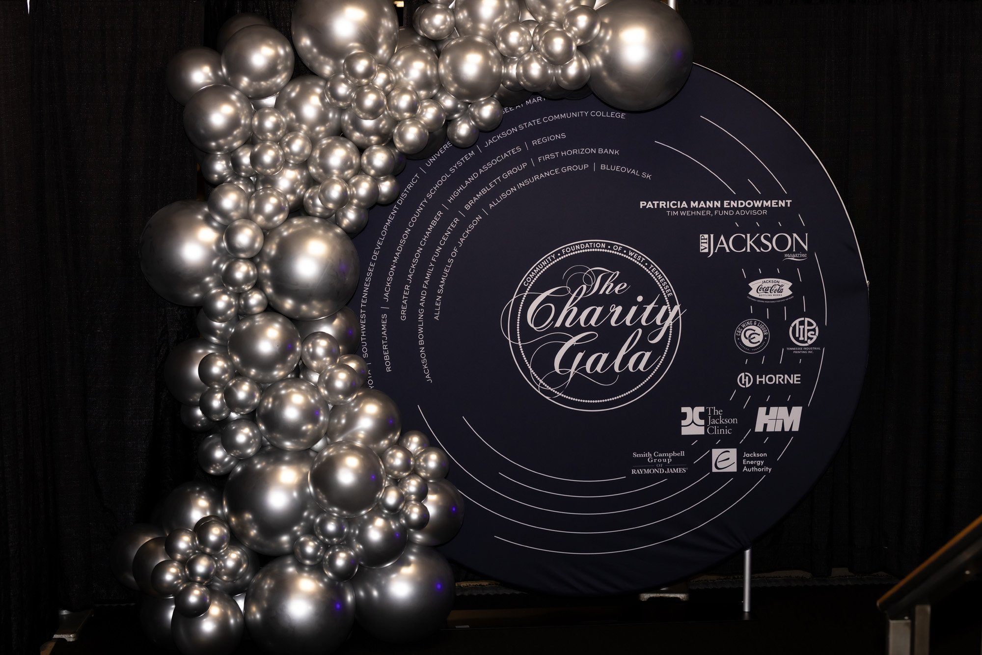 Silver balloon arch surrounding a black circular sign with white text that reads 'The Charity Gala' and lists sponsors and supporters of the event.