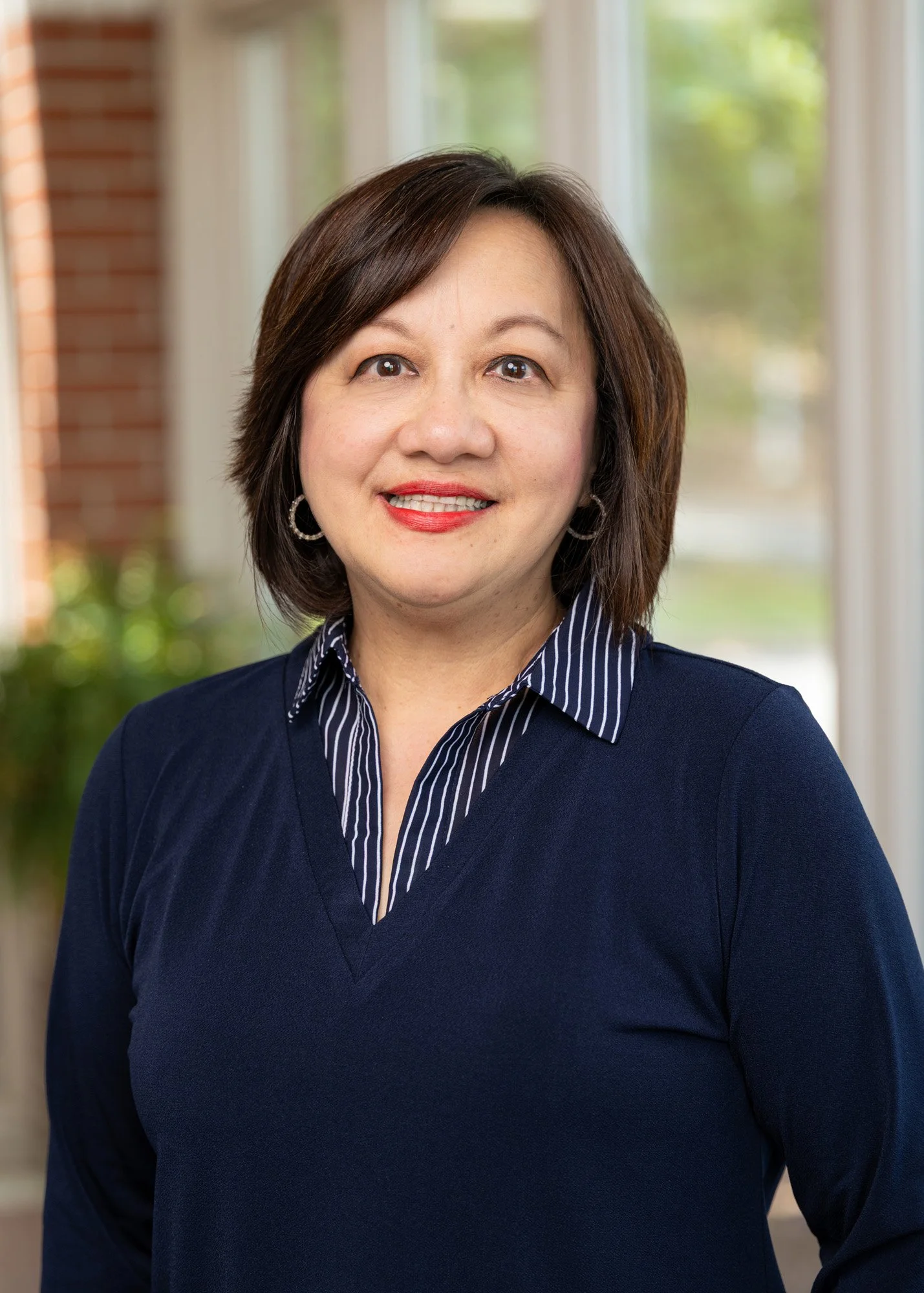 Portrait of a woman with short dark hair, wearing hoop earrings and a navy blue sweater over a striped shirt, standing inside with a blurred background of windows and trees.
