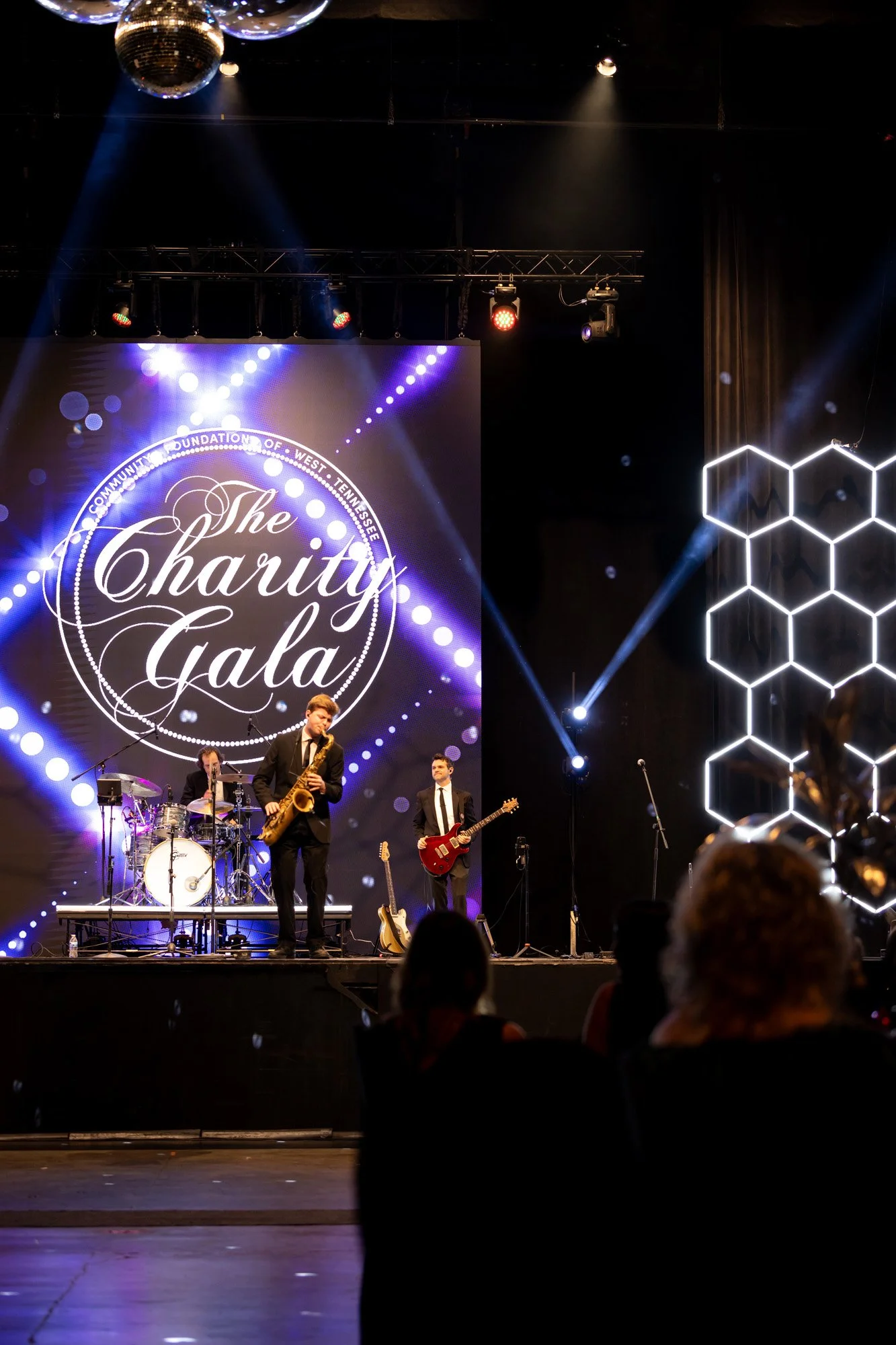 Band performing on stage at a charity gala, with a large screen displaying the event's name and logo, stage lights, and audience members in the foreground.