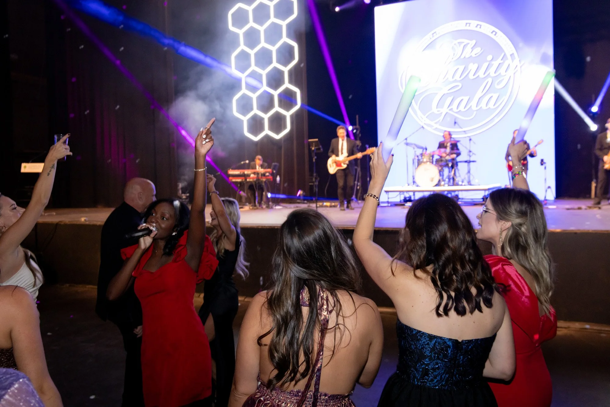People dancing and singing at a Charity Gala with a live band on stage and a large screen behind displaying 'The Charity Gala' logo, with purple and white stage lighting and hexagonal light design.