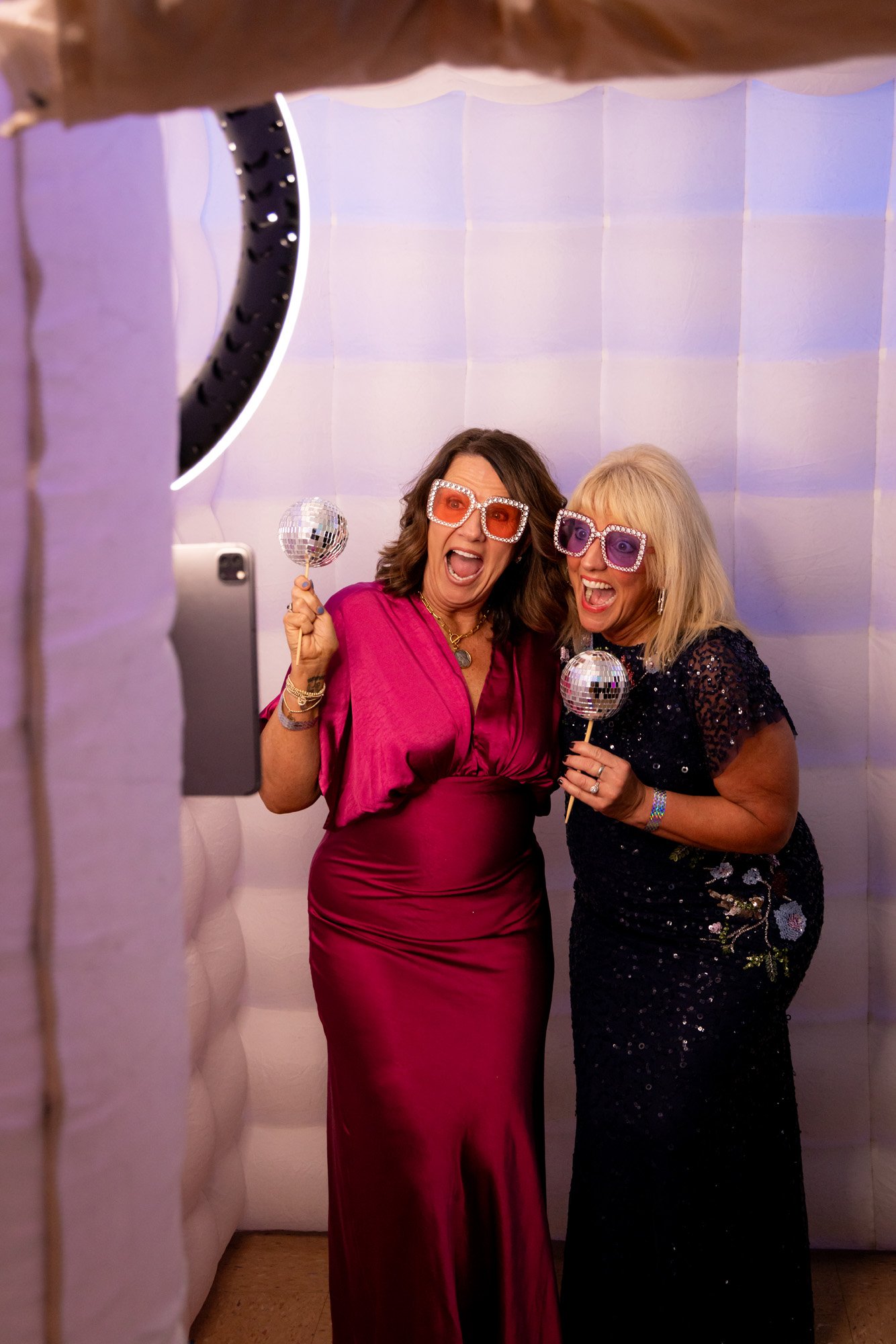 Two women in glamorous dresses and large sunglasses celebrating with miniature disco ball props in a photo booth, smiling