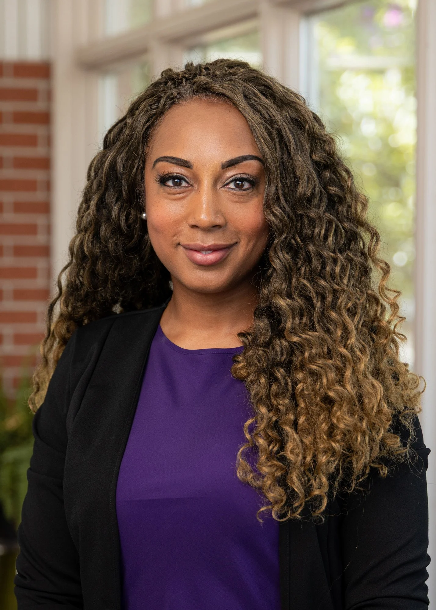 A woman with curly hair wearing a purple top and black blazer, smiling, standing in front of a background with windows and greenery.