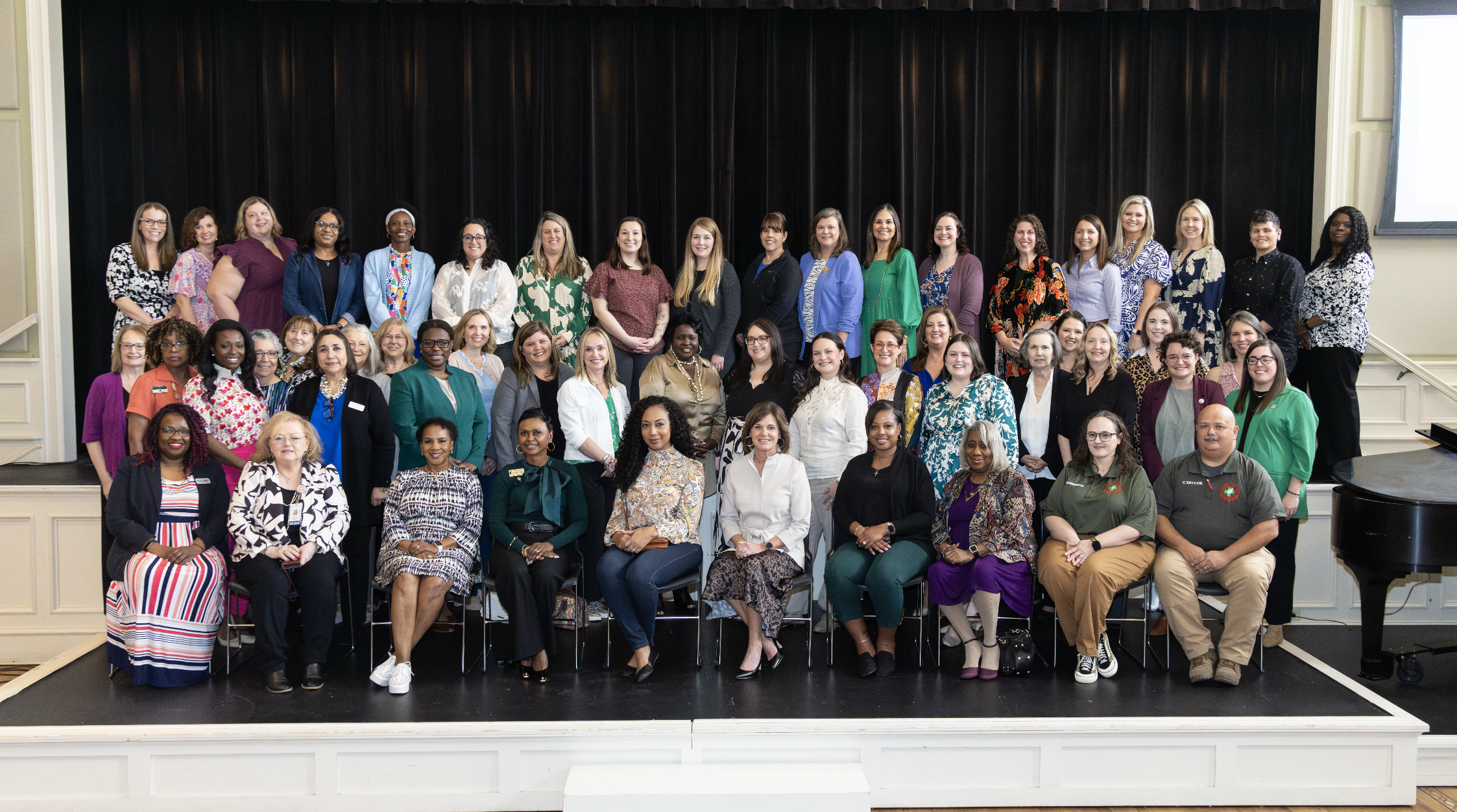 A large group of 100 women and a few men pose on a stage against a dark curtain backdrop. The group is diverse in age and ethnicity, wearing a mix of professional and casual attire, smiling for the photo at an indoor event or conference.