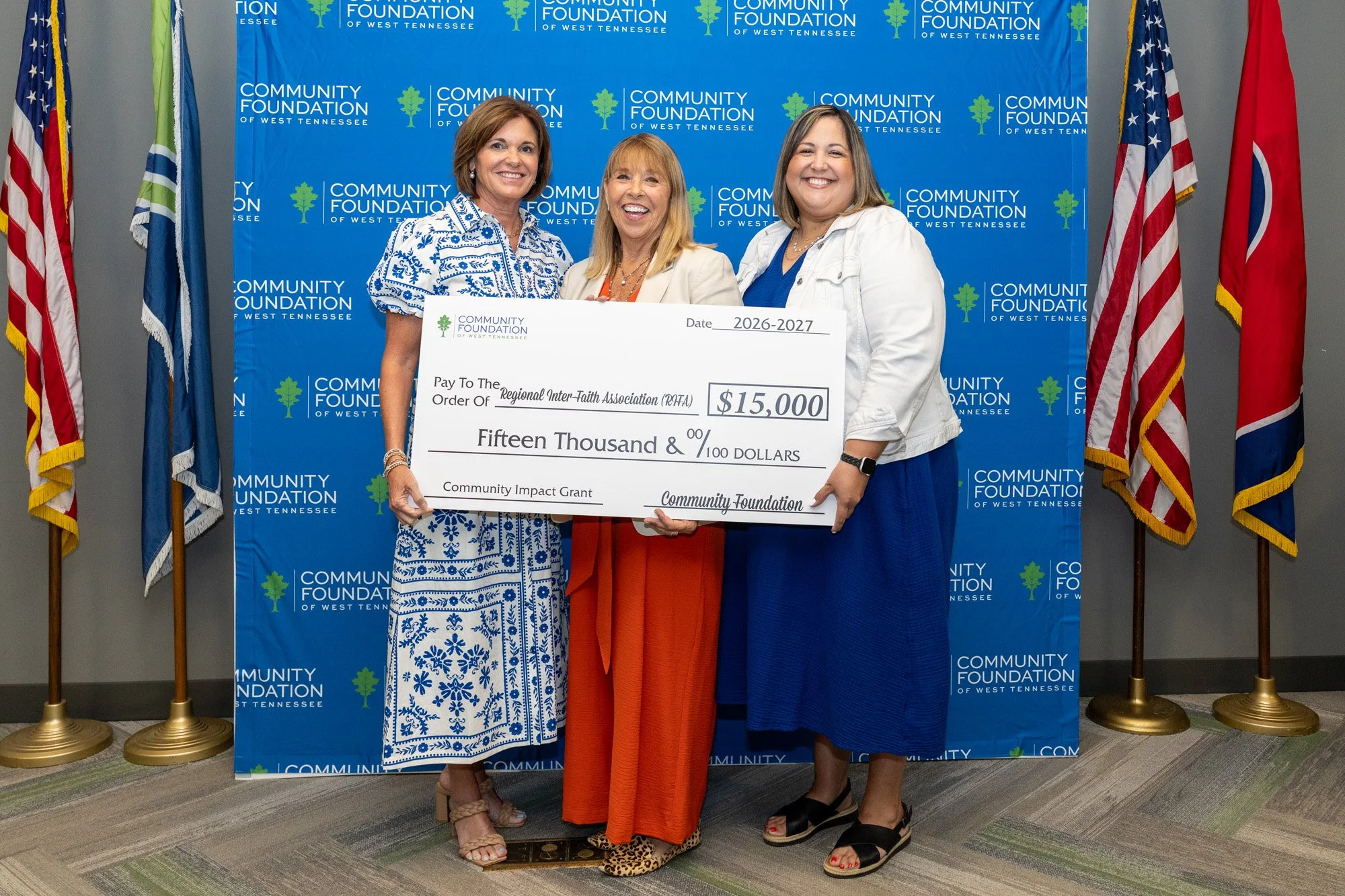 Three women standing side by side holding a large ceremonial check at a community event, with flags and a blue backdrop with green trees and the words 'Community Foundation of West Tennessee' behind them.
