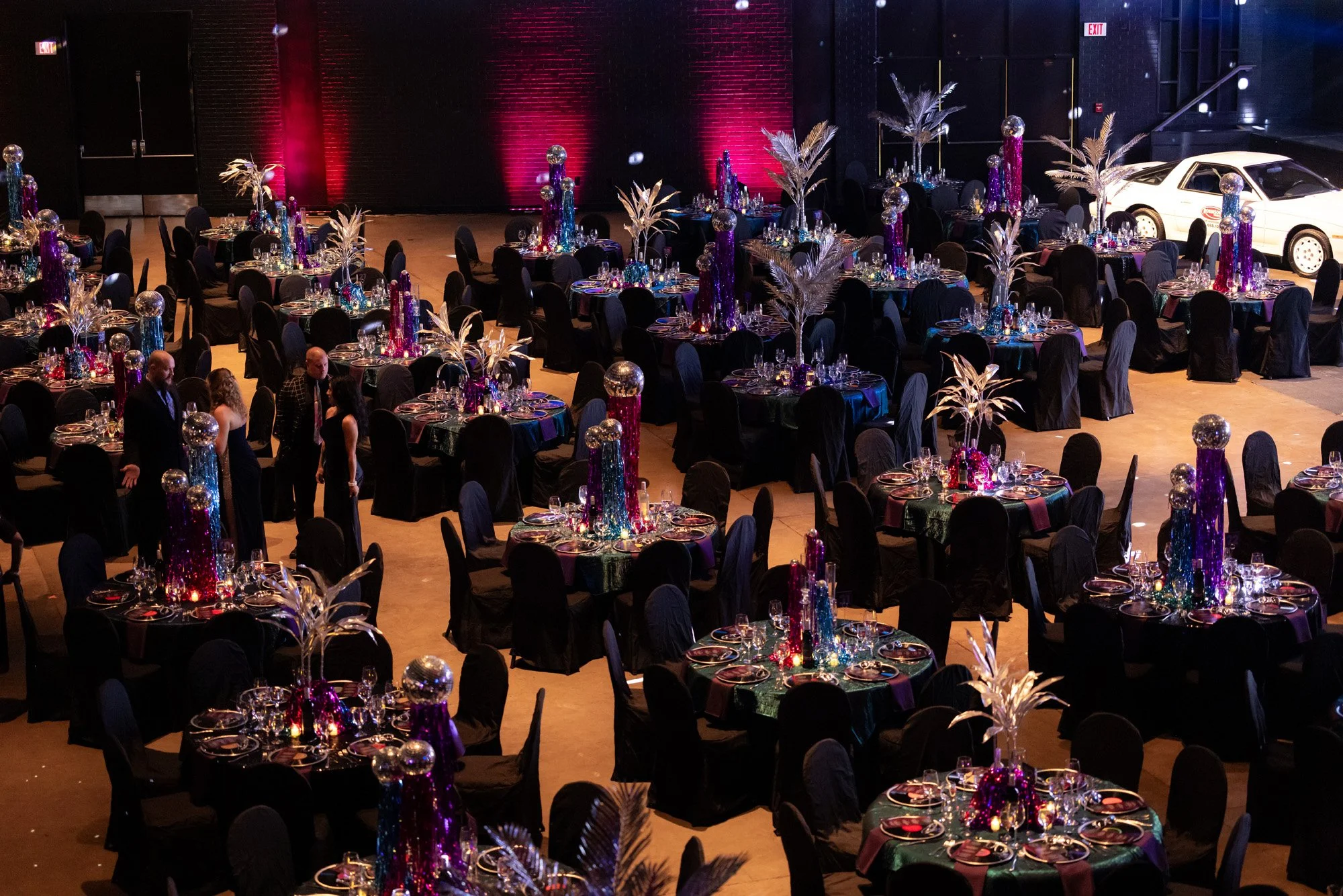 A decorated banquet hall with round tables covered in black tablecloths, decorated with colorful centerpieces featuring tall vases in purple, blue, and pink with metallic accents, set for a formal event.