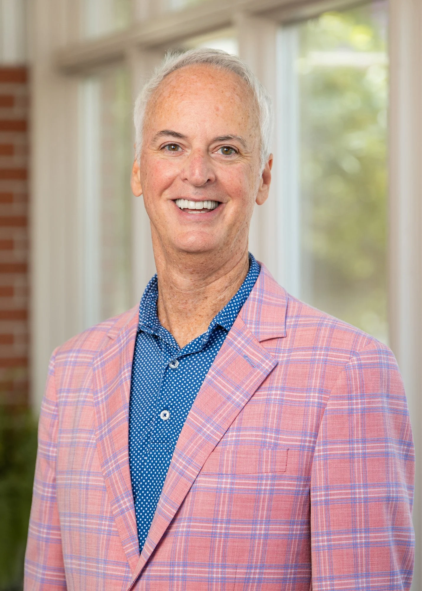 A smiling middle-aged man with gray hair wearing a pink checkered blazer and a blue polka dot shirt, standing in front of windows with natural light.