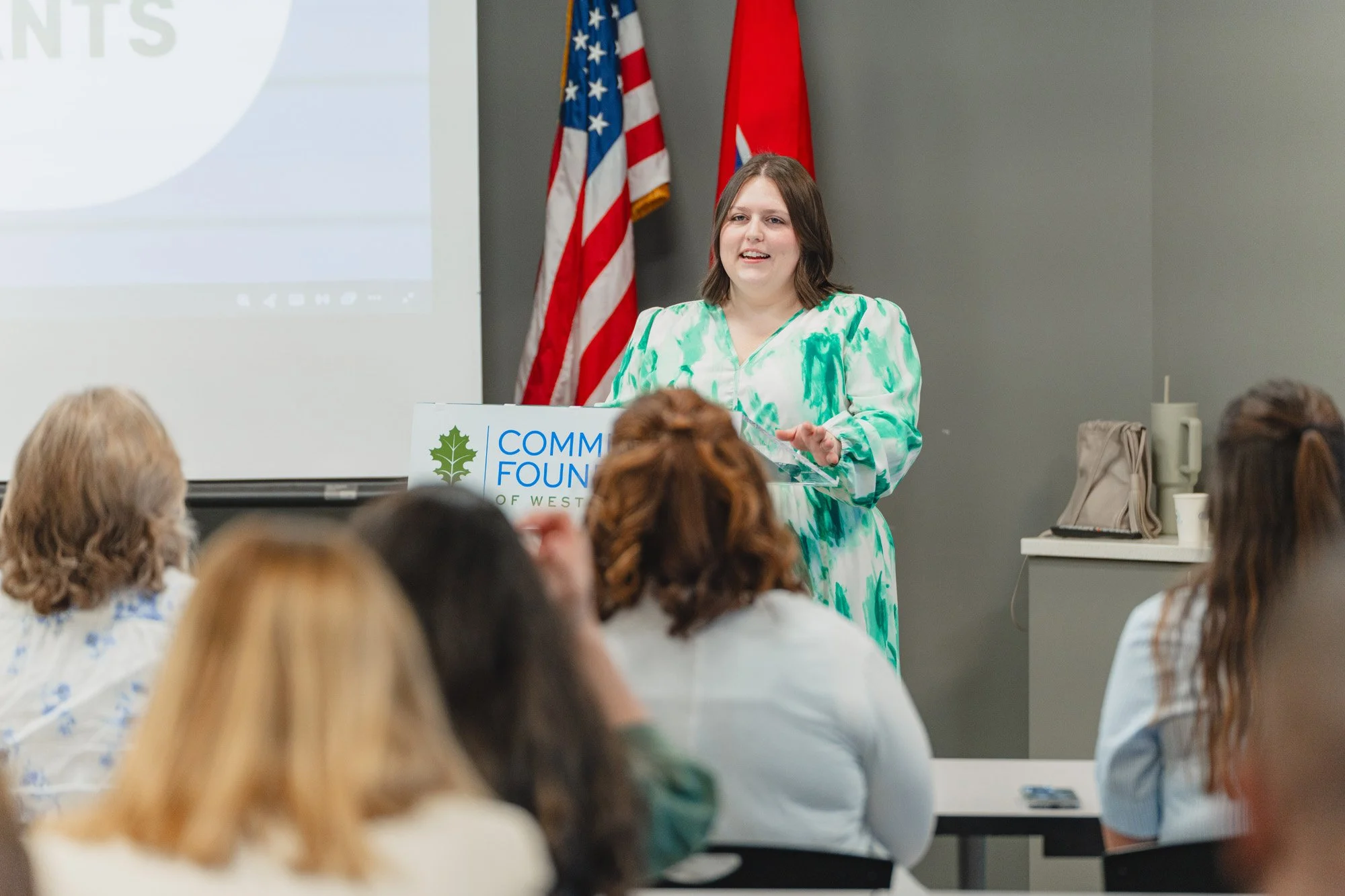 A woman with shoulder-length brown hair stands at a podium giving a presentation to an audience. She is wearing a green and white patterned dress.