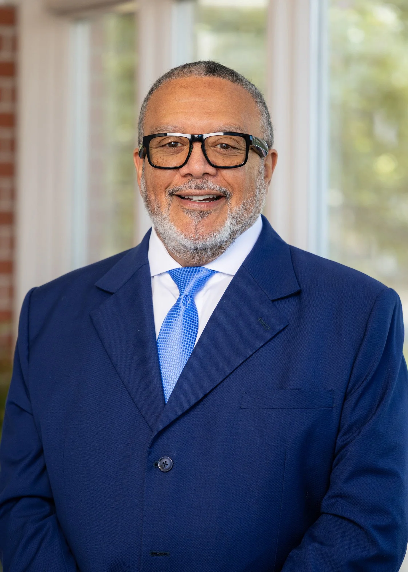 A smiling middle-aged man with glasses, a beard, and short gray hair wearing a blue suit, white shirt, and blue tie, standing in front of a window with blurred greenery outside.