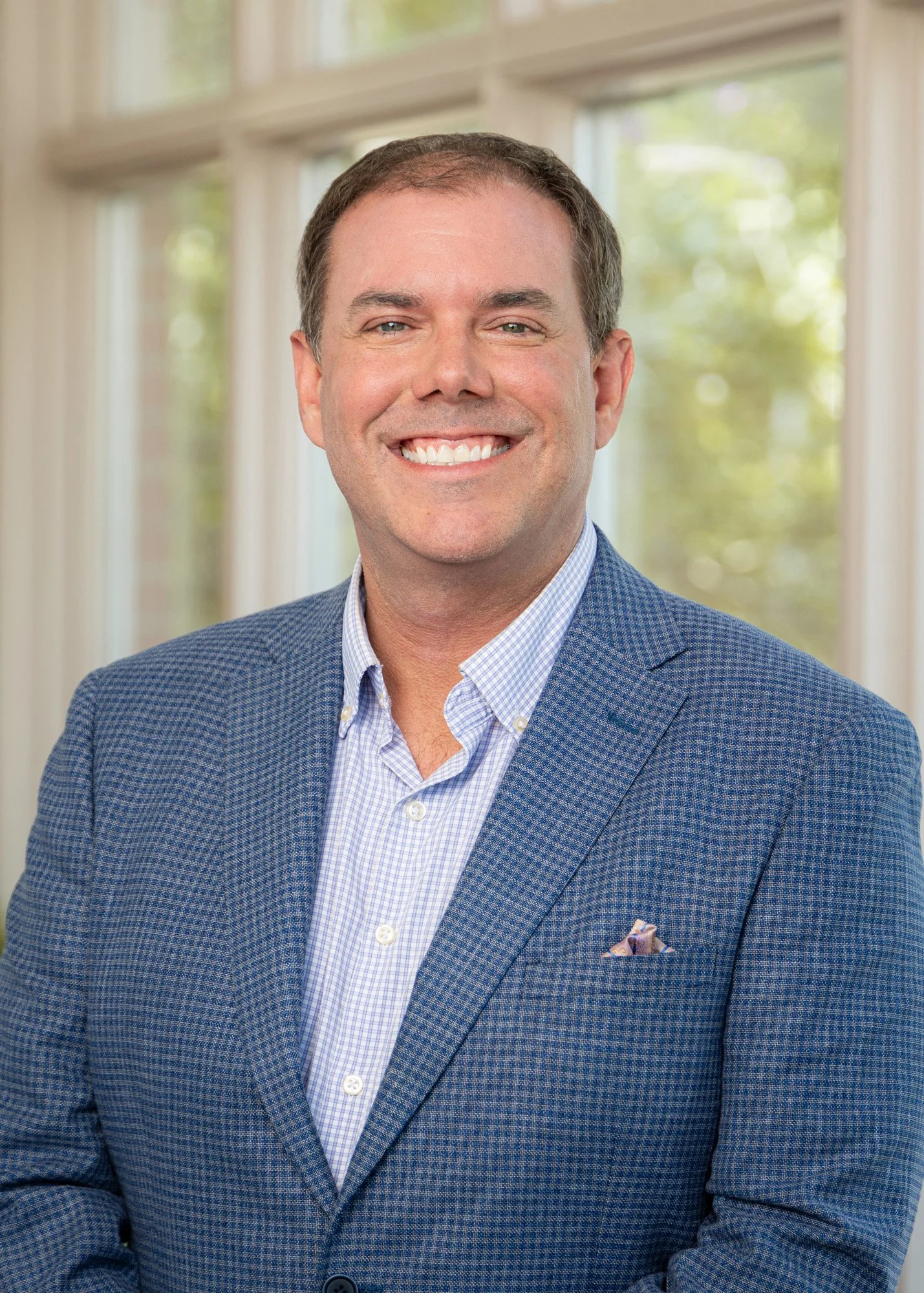 A smiling man with short dark hair, wearing a blue patterned blazer, light blue shirt, and a pocket square. He is standing indoors with large windows and greenery in the background.