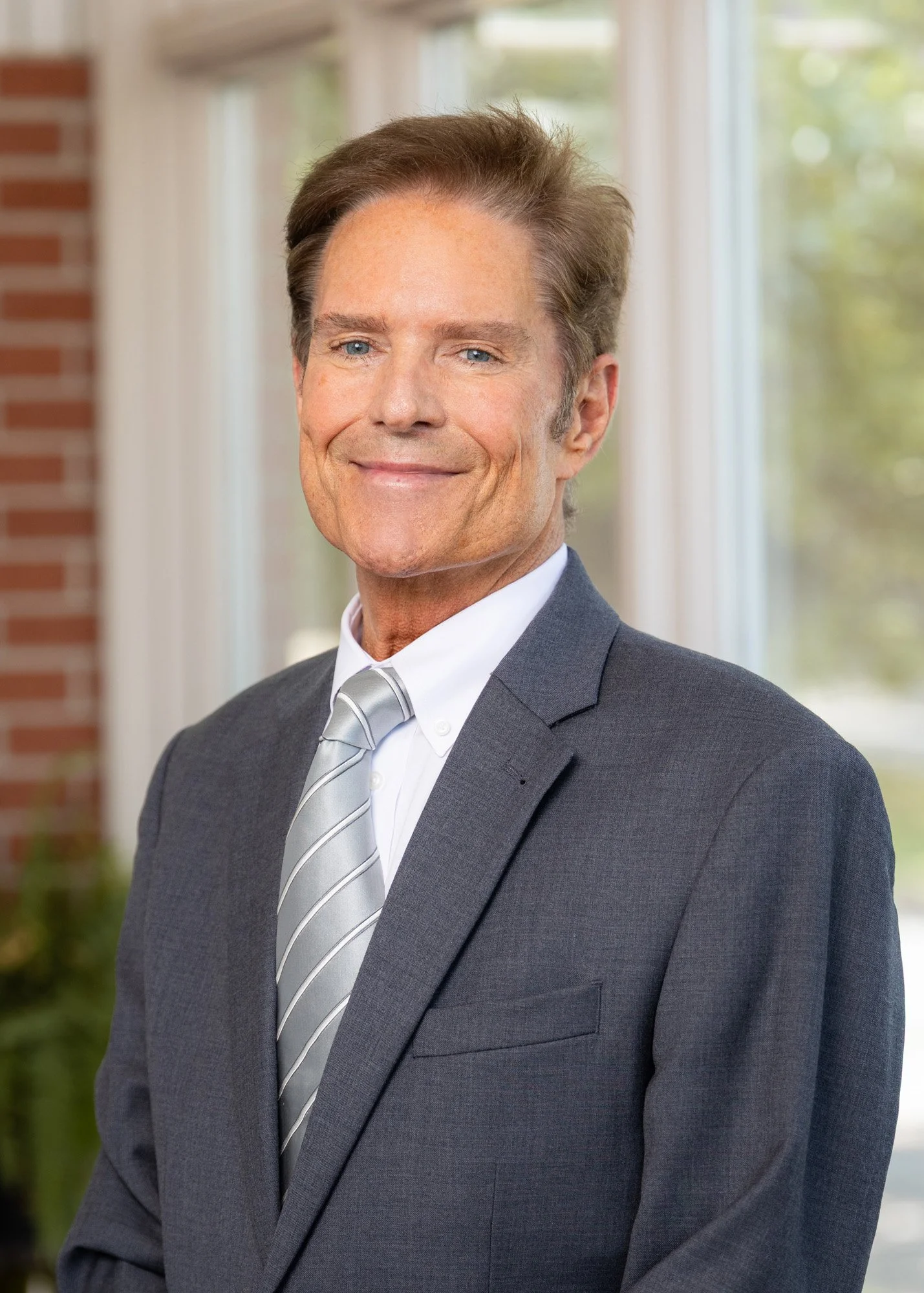 A middle-aged man with blue eyes and brown hair in a gray suit, white shirt, and striped tie, smiling softly, standing indoors with blurred windows and brick wall in the background.