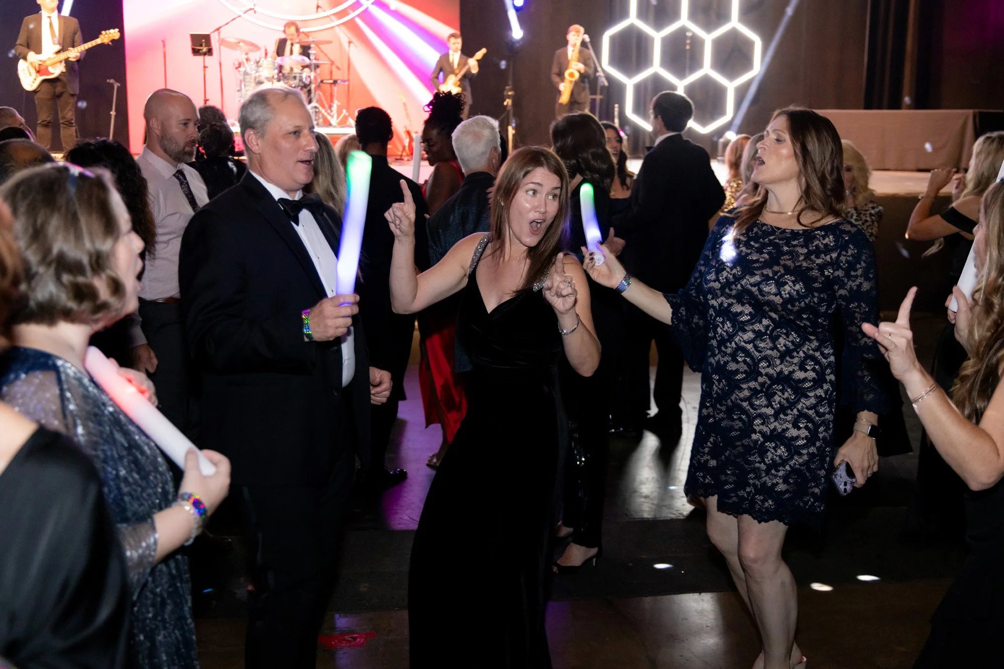 People dancing and socializing at a lively party with a live band playing on stage in the background, colorful lighting, and hexagonal light decor.