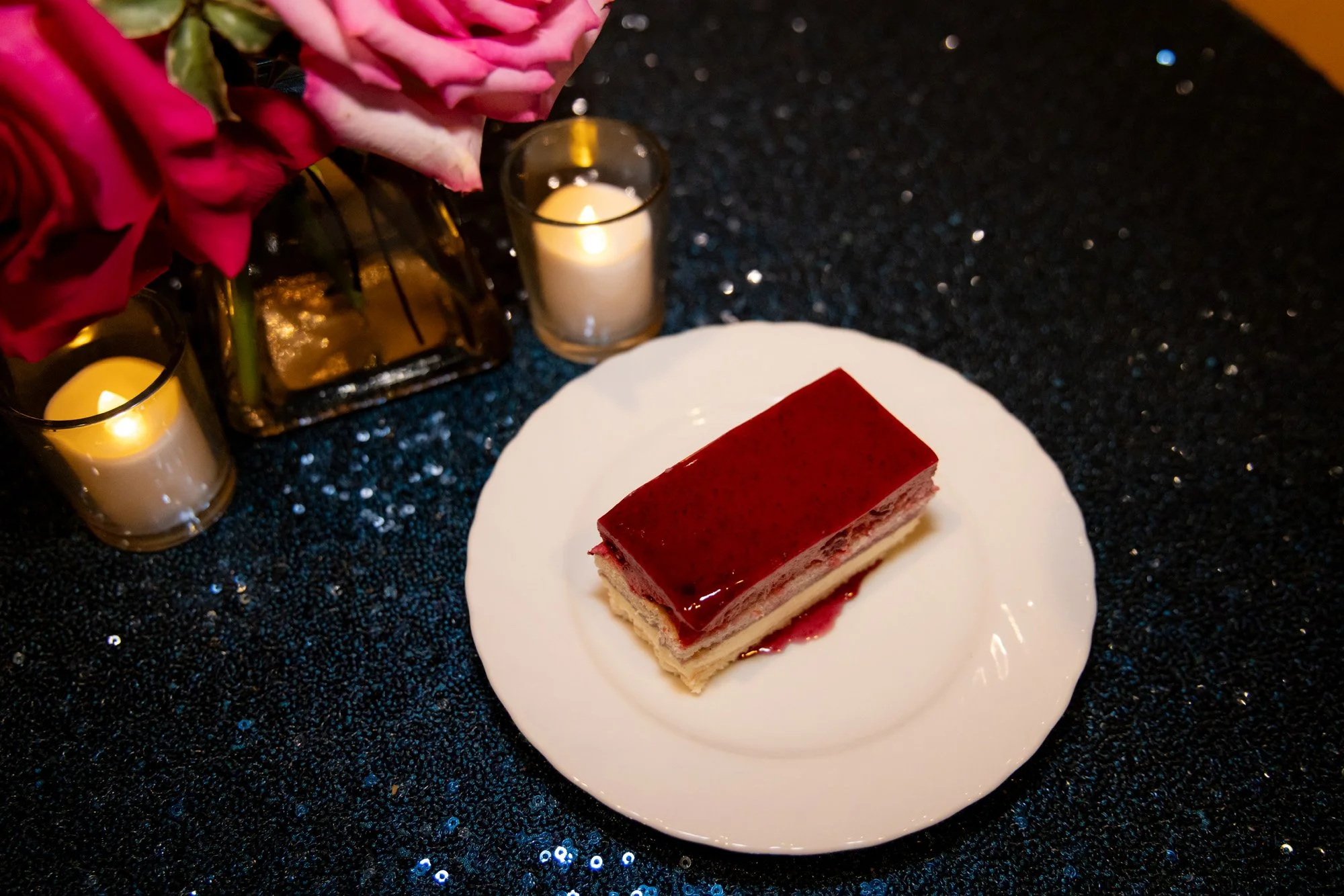 A slice of layered cake with a red glaze on top, served on a white plate, on a black sparkly tablecloth, with pink roses and lit candles in the background.
