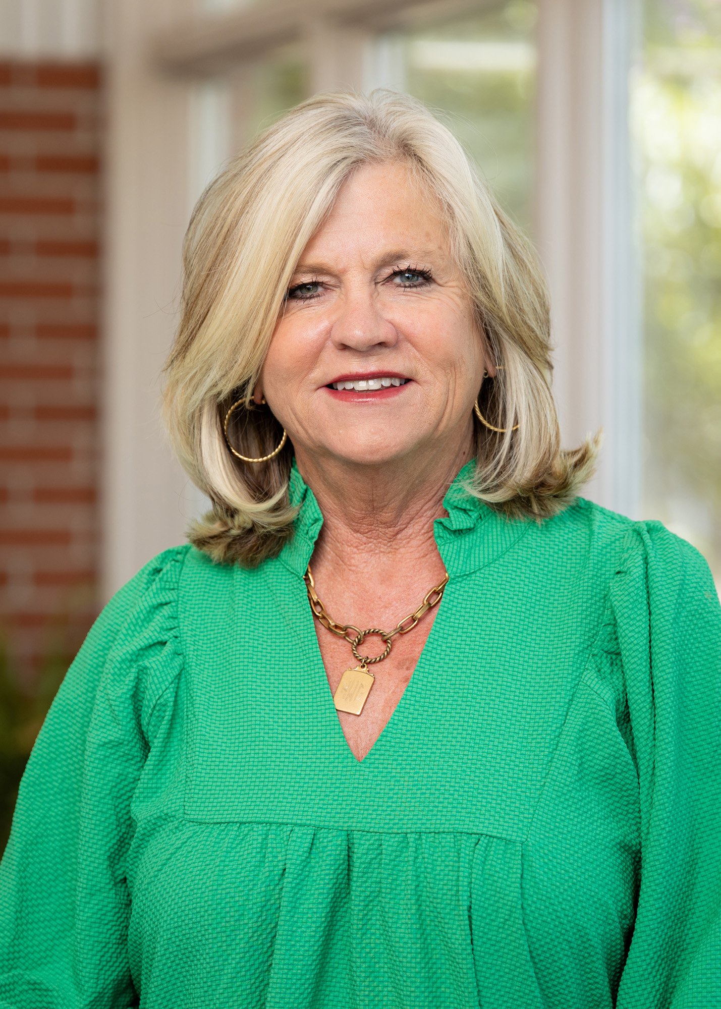 A smiling middle-aged woman with shoulder-length blonde hair, wearing a green textured blouse and gold jewelry. She is indoors with a brick wall and large window with greenery outside in the background.