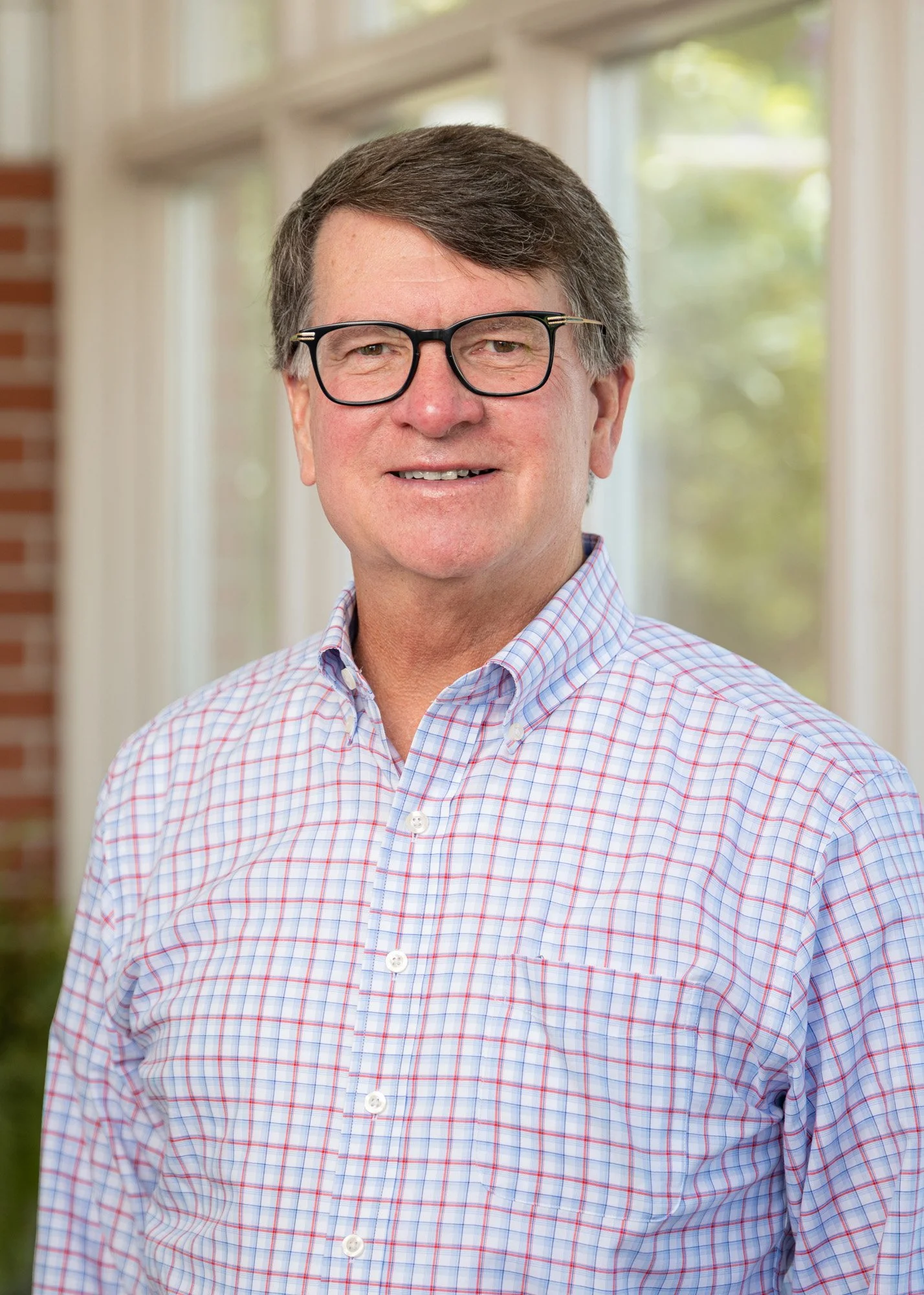 A middle-aged man with dark hair, wearing glasses and a checkered dress shirt, standing in front of large windows with green trees outside.