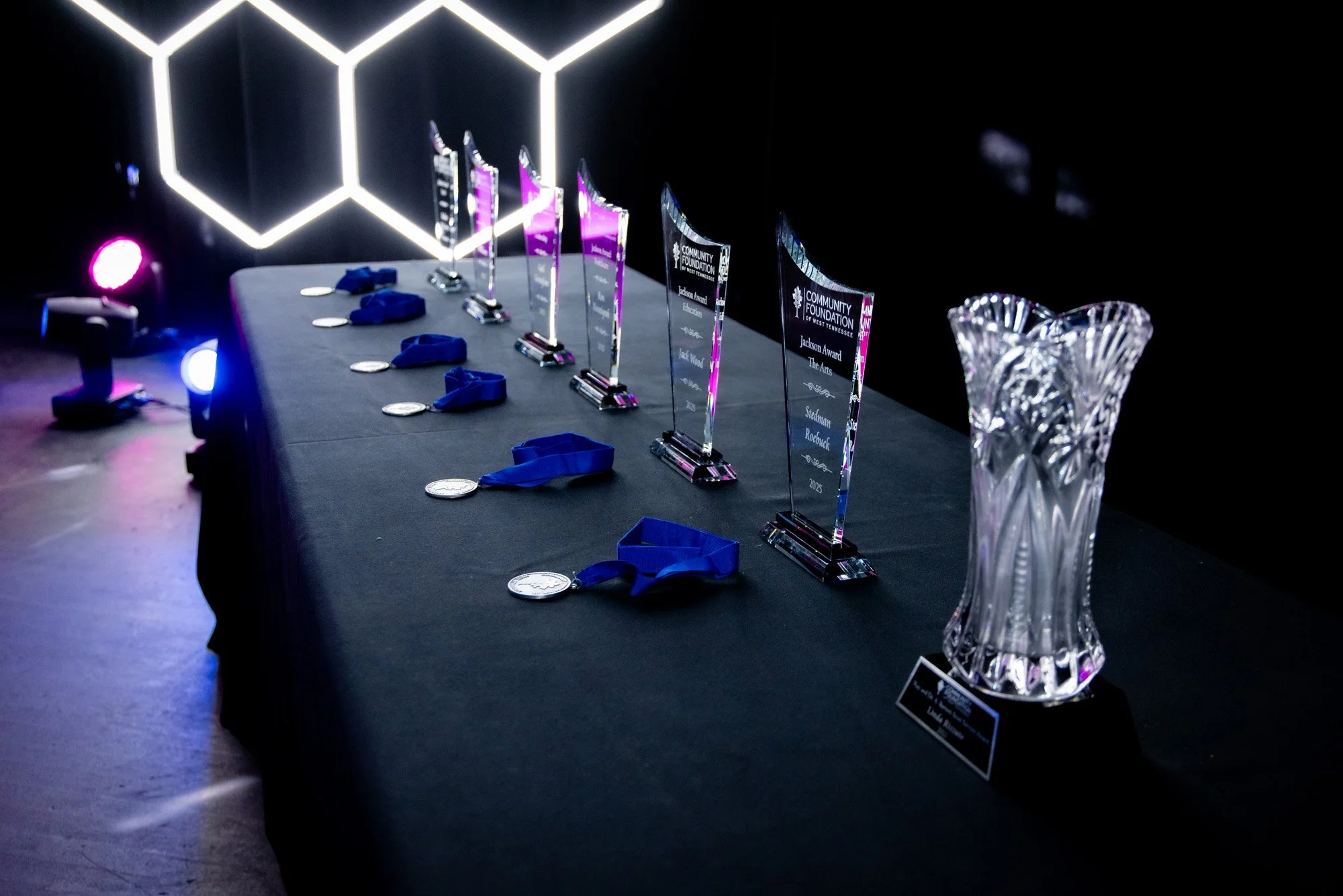 Awards table with several glass trophies and medals, decorated with blue ribbons, against a dark background with a geometric light installation and a crystal vase on the right.