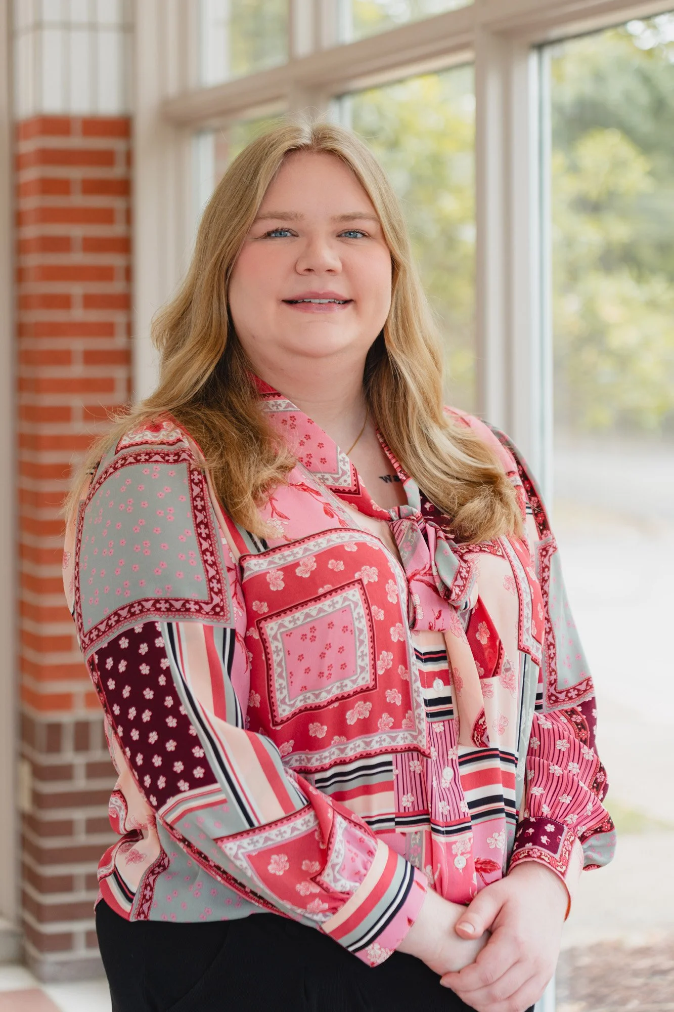 A woman with long blonde hair and blue eyes standing near a window with a brick wall in the background, wearing a colorful, patterned blouse with red, pink, gray, and white colors.