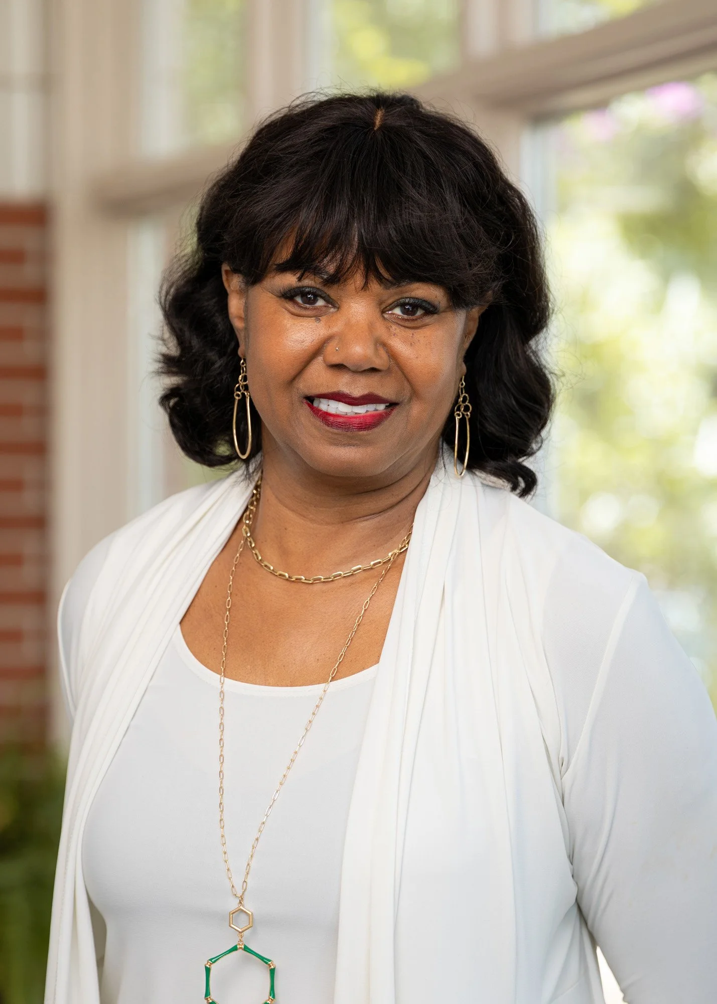 A woman with dark hair styled in soft curls, wearing a white top with a white blaze and gold jewelry, smiling and standing in front of a window with greenery outside.
