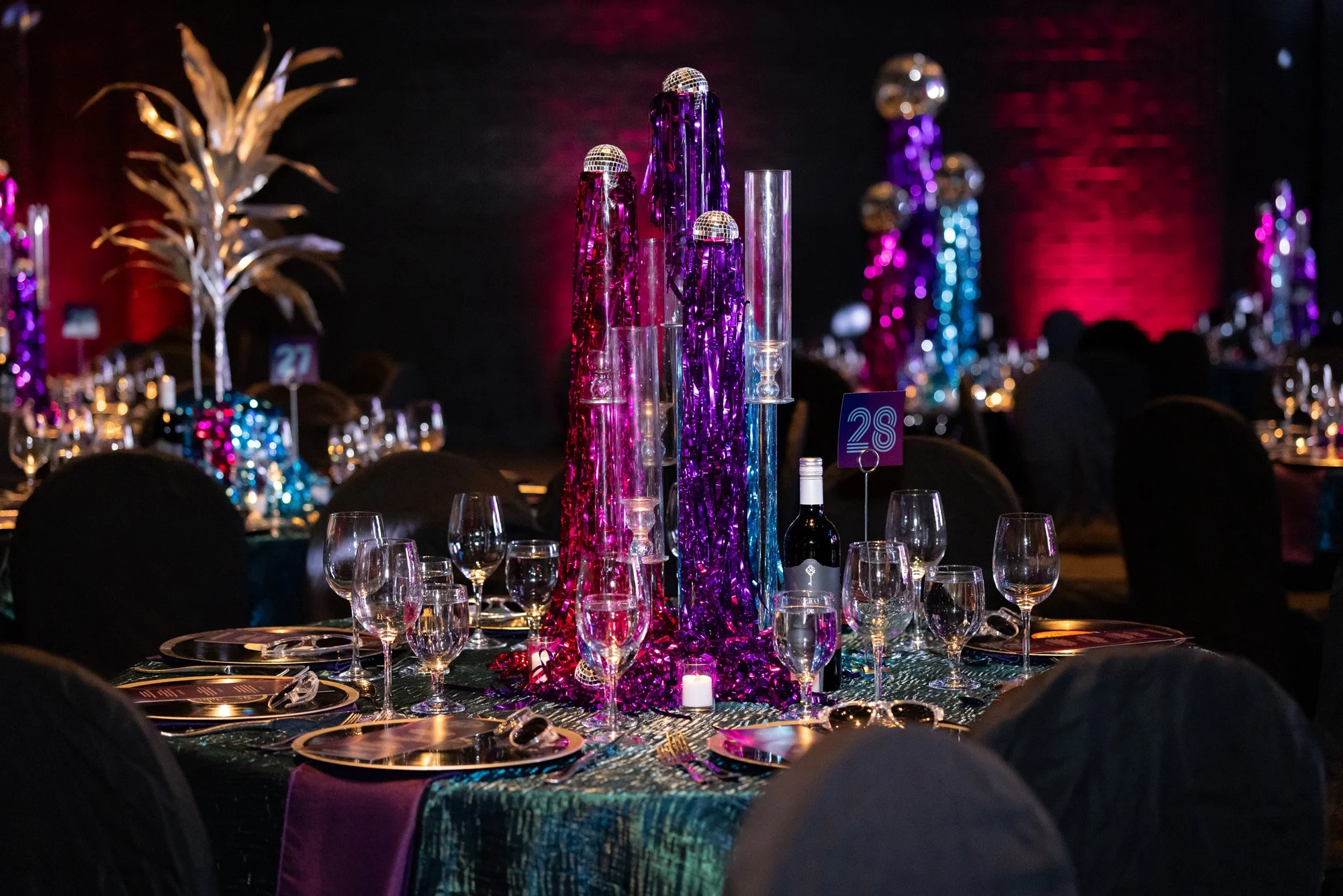 Elegant banquet table with tall colorful centerpieces, wine glasses, plates, and a bottle of wine in a dimly lit event venue.
