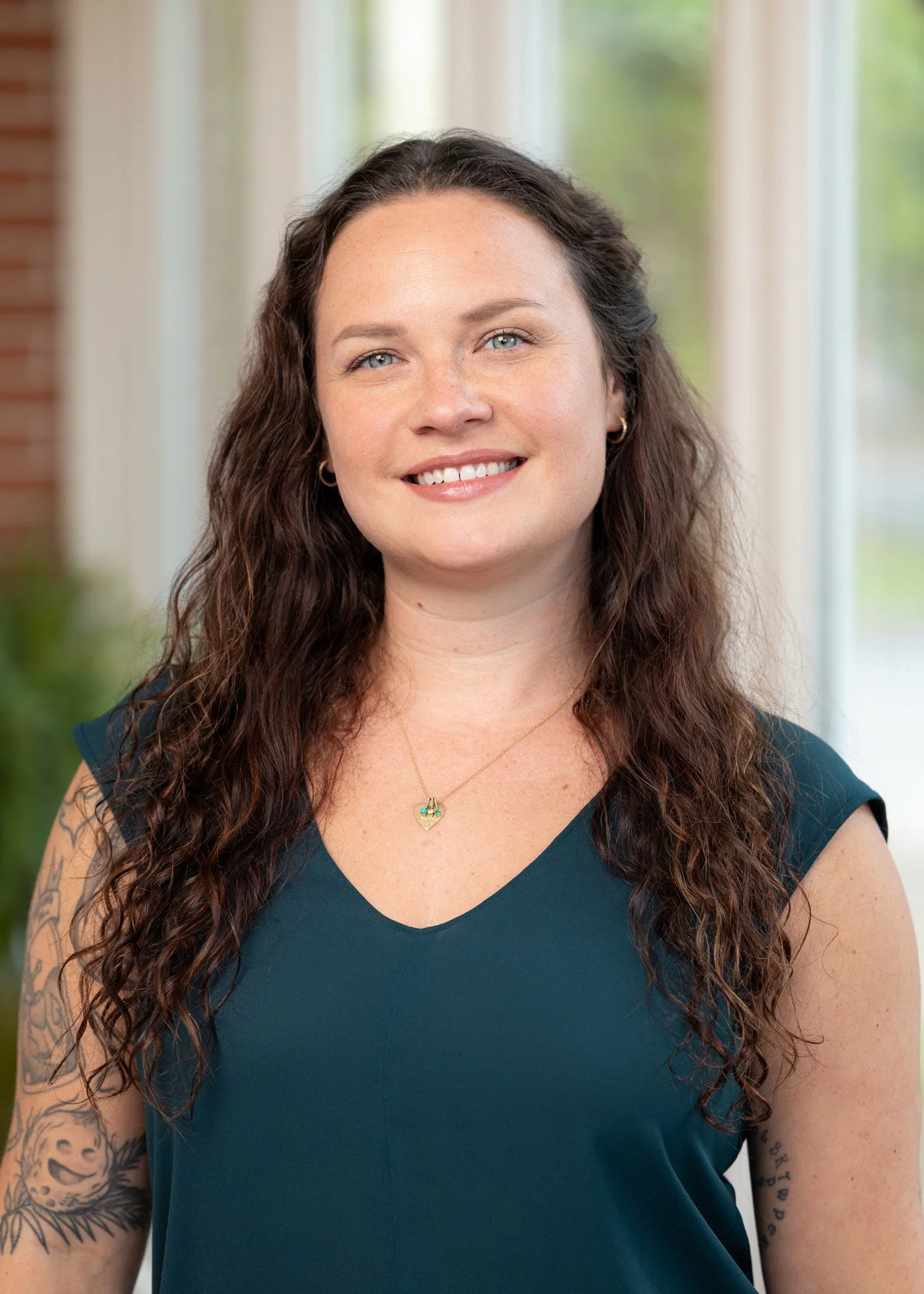 Woman with long, curly brown hair and blue eyes, smiling, wearing a dark blue top with gold jewelry and tattoos on her arms, standing indoors with a blurred background of windows and greenery.