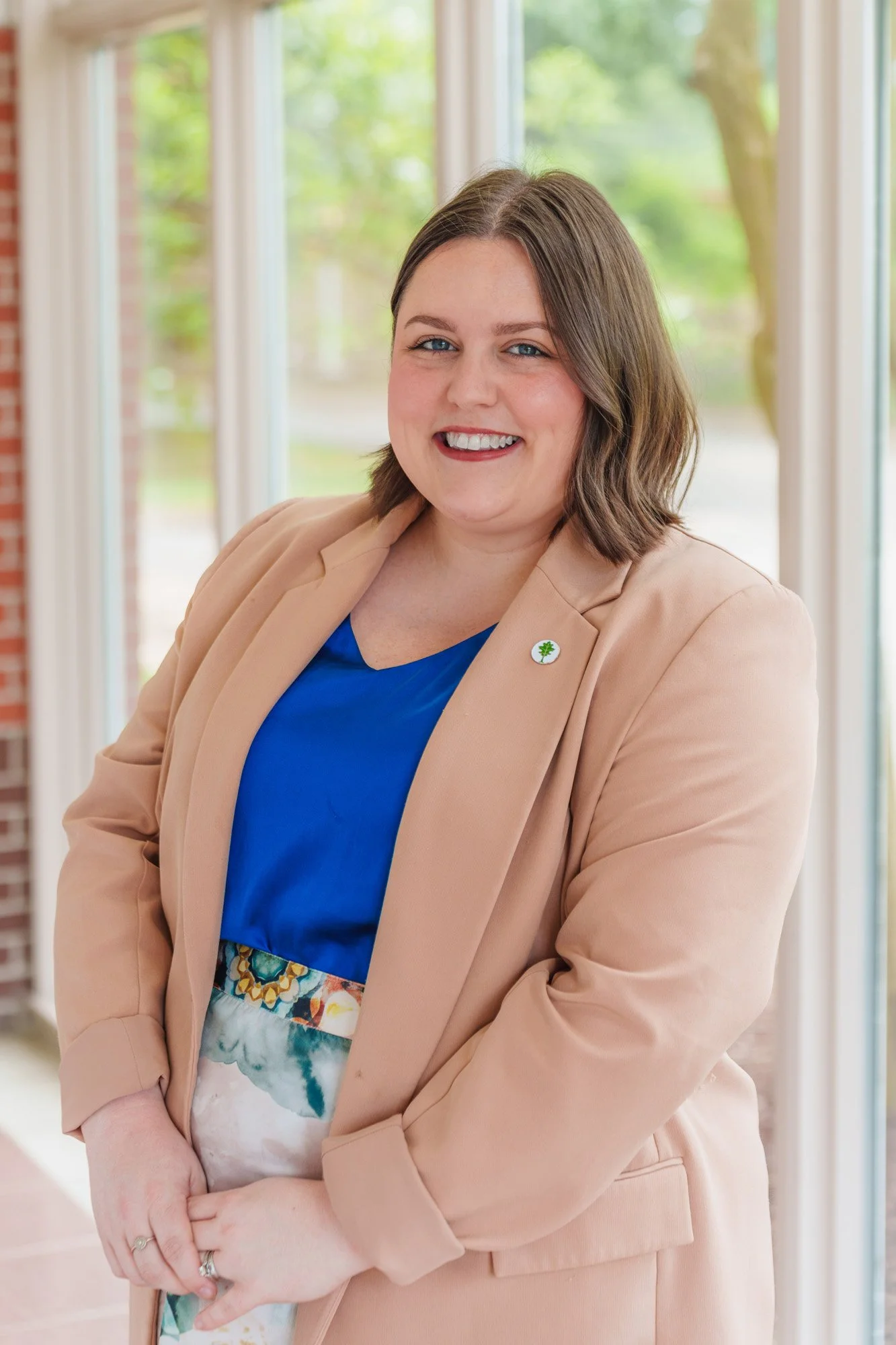 A woman with shoulder-length brown hair, blue eyes, and a tan blazer smiling in front of large windows with green trees outside.