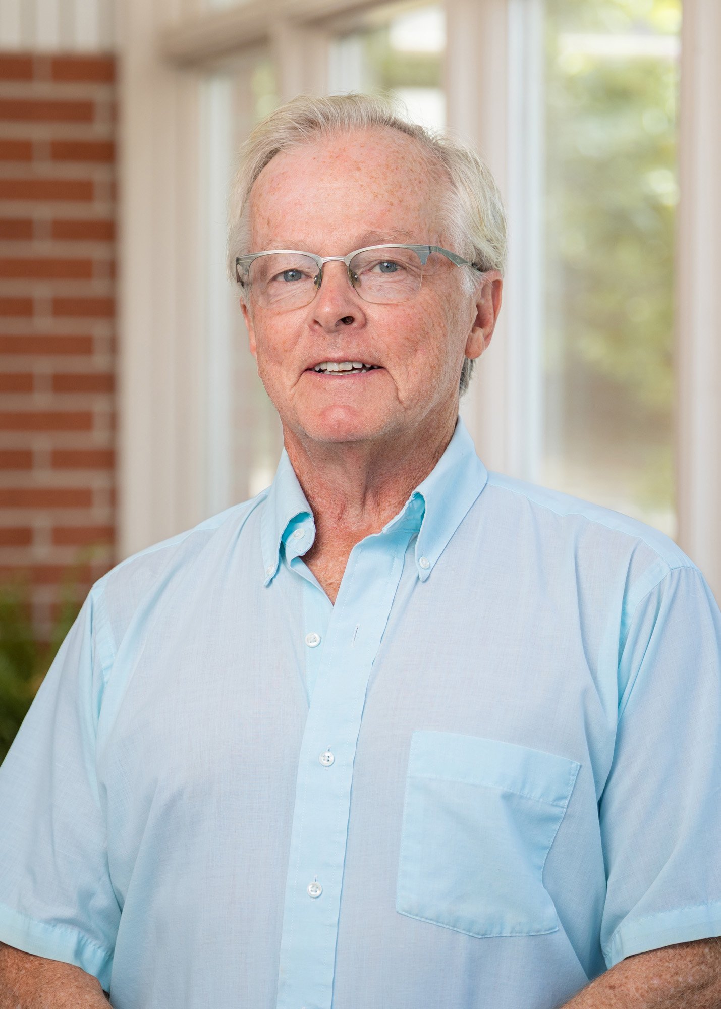 Older man with glasses in a light blue shirt standing indoors near windows and brick wall, smiling.