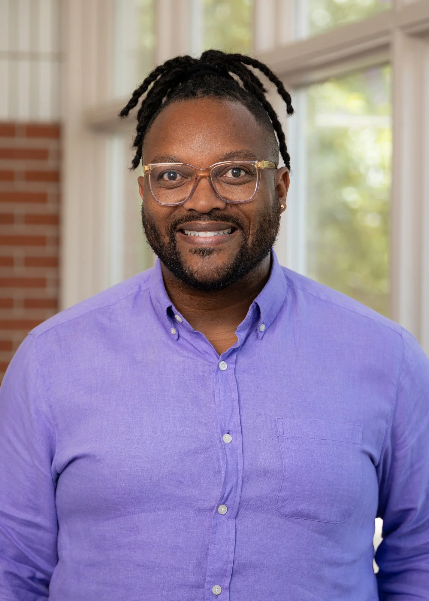 A man with dark skin, dreadlocked hair, glasses, a beard, and wearing a purple button-up shirt is smiling in a brightly lit room with large windows and a brick wall.
